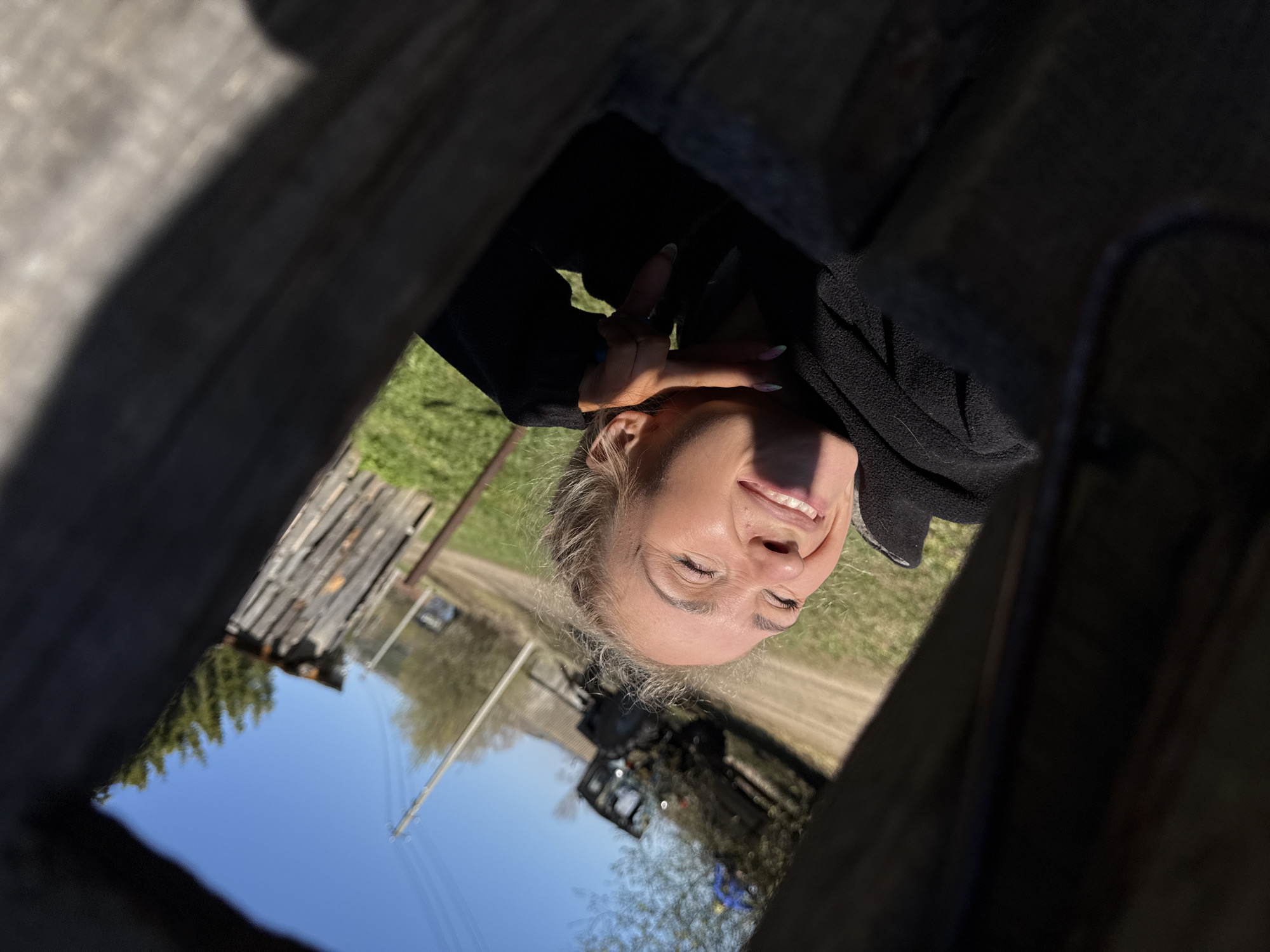 The image shows a smiling woman in Nizhnyaya Chermoda, Russia, as seen through a wooden frame. The background includes a dirt road, a utility pole, a tractor, and some buildings. The sky is blue. The woman is wearing a dark jacket and has blonde hair. She is holding up her hand near her face.