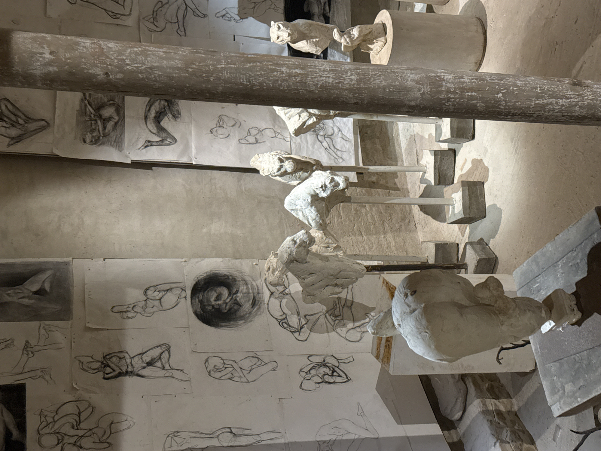 The image depicts an art studio space in Stellenbosch, South Africa. The walls are covered in charcoal sketches of human figures in various poses. Sculptures of human torsos, heads, and full figures in plaster are displayed on individual stands. A horizontal beam bisects the image's foreground. The overall aesthetic suggests a teaching or working studio.