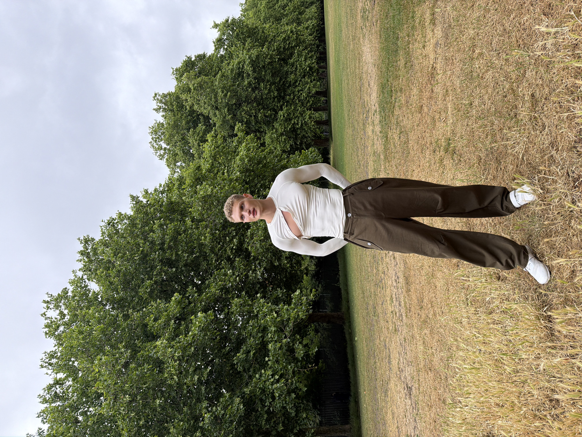 A full-length portrait shows a young person standing outdoors in a park or large grassy area. The subject, who appears male, has short blonde hair and fair skin. He is centrally positioned in the frame, facing the camera with a neutral expression and a confident pose, with his hands placed on his hips. He is dressed in a fitted, long-sleeved, off-white or cream top featuring a distinctive wrap-around or cut-out detail on the chest. This is paired with dark olive green or brown loose-fitting cargo-style trousers and white sneakers.
The background features an expansive field of grass, which displays variations in color from green to dry golden-brown patches, with some taller wild grass visible, particularly in the foreground. Behind the subject, a subtle dark fence runs along the edge of the grassy area, separating it from a dense line of tall, leafy green trees that dominate the middle ground. The sky above is light grey and uniformly overcast, indicating a cloudy day with soft, diffused lighting. The overall scene suggests daytime, likely morning or afternoon, in a spacious outdoor environment, consistent with a large park in London, United Kingdom. There is no visible text in the image.