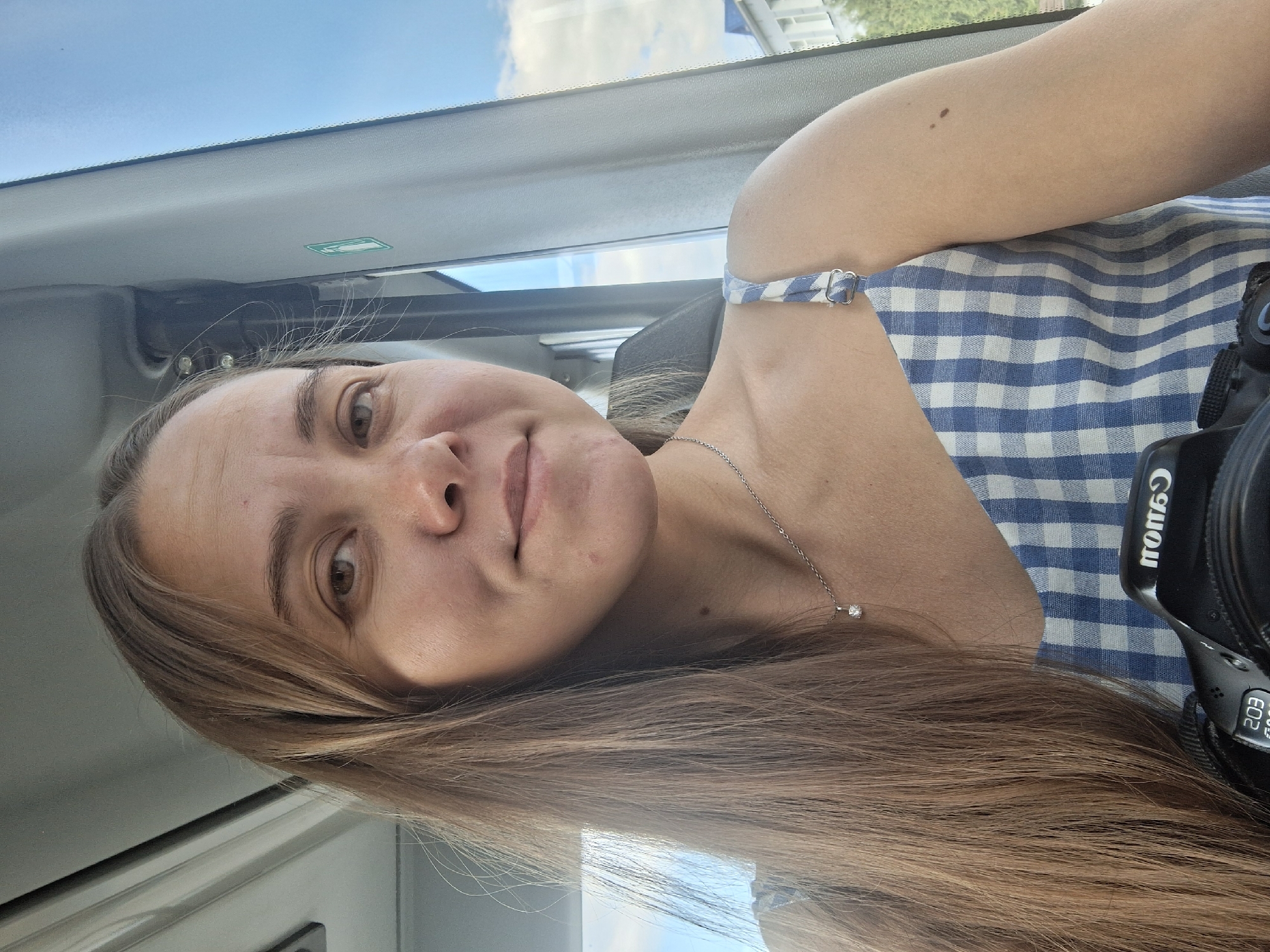 A female individual, appearing to be in her late 20s or early 30s, is centered in a close-up, chest-up self-portrait. She has long, light brown hair, parted to her left, that cascades over her shoulders. Her brown eyes are open and directed at the viewer, and she exhibits a slight, closed-mouth smile. Visible forehead lines are present. She is adorned with a thin silver chain necklace featuring a small, clear gemstone pendant. She wears a light blue and white gingham-patterned top with a thin shoulder strap visible on her right. A small dark mole is visible on her right upper arm and another on her lower left neck. She is holding a black Canon EOS 20D DSLR camera, which is partially visible at the bottom of the frame.
The individual is seated inside what appears to be a public transport vehicle, such as a bus or train. The interior features a light-colored ceiling and window frames, with a gray seat headrest partially visible behind her. A small green square sign with a white pictogram is affixed to the right window frame. Outside the window, a clear blue sky with scattered white clouds is visible, alongside distant light-colored buildings and green trees, indicating an urban or suburban setting. The location is the city of Chaikovsky, Russia.