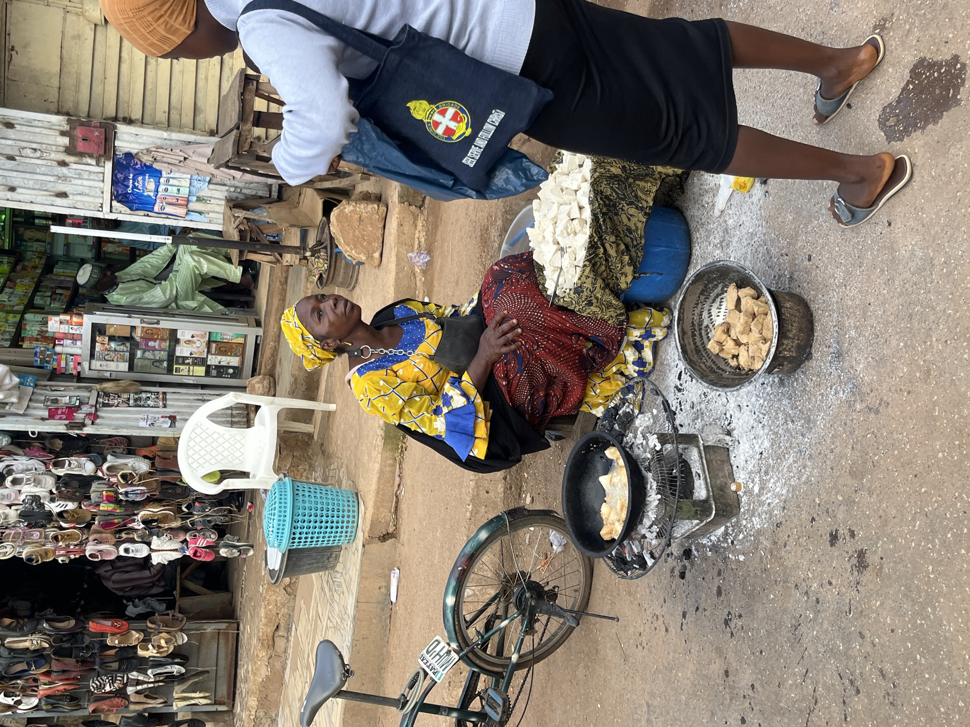 The image depicts an outdoor marketplace scene in Bukuru, Nigeria. A woman wearing a bright yellow and blue patterned dress and a yellow headscarf sits on the ground. She is surrounded by various goods, including a blue plastic tub filled with cubed white food items, a pan of fried food over a charcoal stove, and a metal bowl of fried food. To her right, another woman, wearing a white top and a black skirt with an orange headscarf, stands and holds a dark blue tote bag with a circular emblem and text. In the background, a shoe stall is visible, displaying a variety of footwear hanging from racks. Two men are partially visible inside the stall; one is seated and wearing a light green traditional outfit and cap, while the other stands behind him. A white plastic chair is also present near the shoe stall. A bicycle is parked to the left of the scene. The ground is paved with concrete, and scattered debris, including ash from the stove, is visible. The overall environment suggests a bustling street market setting.