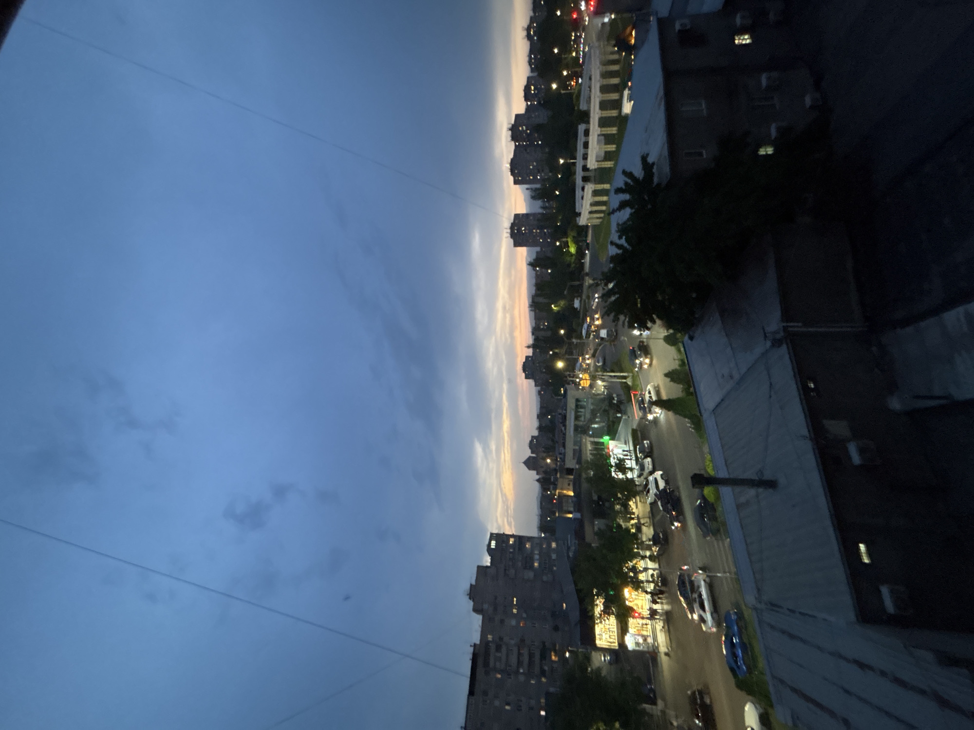 The image captures a cityscape at dusk, likely in Yerevan, Armenia, based on architectural and urban planning characteristics. The view is from an elevated position, possibly a high-rise building. The sky transitions from deep blue to pale orange near the horizon, suggesting either sunrise or sunset. Below, streets are illuminated by streetlights and vehicle headlights. Buildings of varying heights and architectural styles are visible, including Soviet-era apartment blocks. There is moderate traffic. Some buildings have visible windows illuminated from within. Trees and green spaces are sparsely distributed. Overhead electrical wires are visible across the sky.