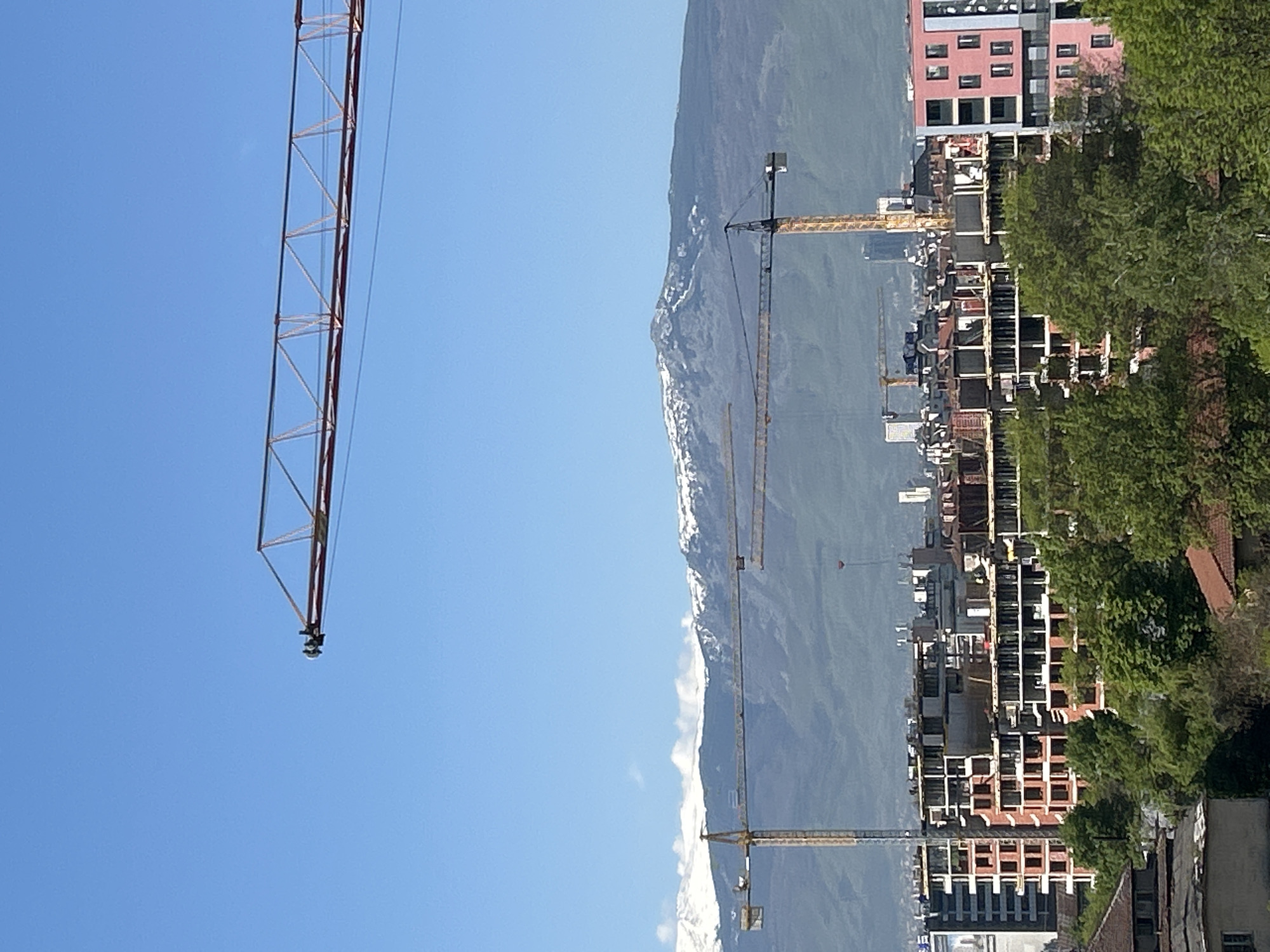 The image shows a cityscape with mountains in the background. Several construction cranes are visible, suggesting ongoing building activity. The buildings are a mix of completed structures and those still under construction. There's a significant amount of greenery in the foreground and middle ground, indicating the presence of trees and possibly parks. The weather appears to be clear with a blue sky. This is near the city of Sofia, Bulgaria.