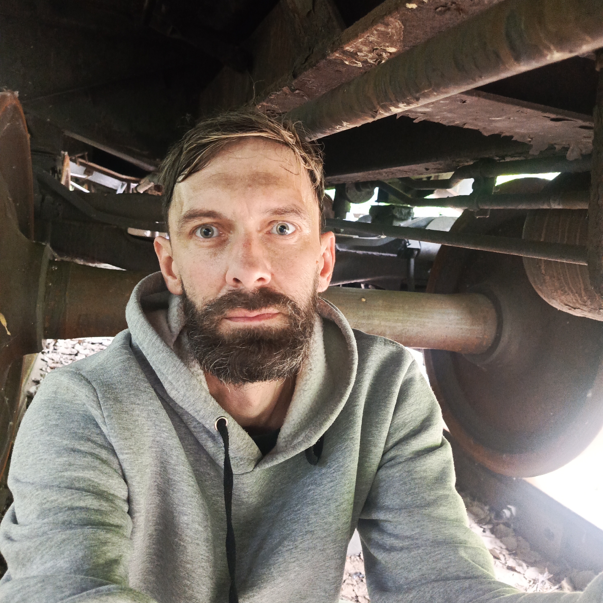 A selfie taken from a low angle, looking up at a man from under a rusty train car. The man, who appears to be in his late 30s or early 40s, has fair skin, light brown hair, and a full beard. He is wearing a grey hooded sweatshirt and is looking directly at the camera with a surprised expression. His eyes are wide open, and his mouth is slightly agape. The train car above him is made of dark, weathered metal, with visible rust and debris. The wheels and undercarriage of the train are also visible, with various metal components and a large, dark-colored wheel. The ground beneath the train appears to be covered in gravel and dirt. The lighting suggests an outdoor environment, possibly during the day. The overall impression is one of an abandoned or disused industrial setting. The specific location in Chaikovsky, Russia, is not identifiable from the visual elements in the image.