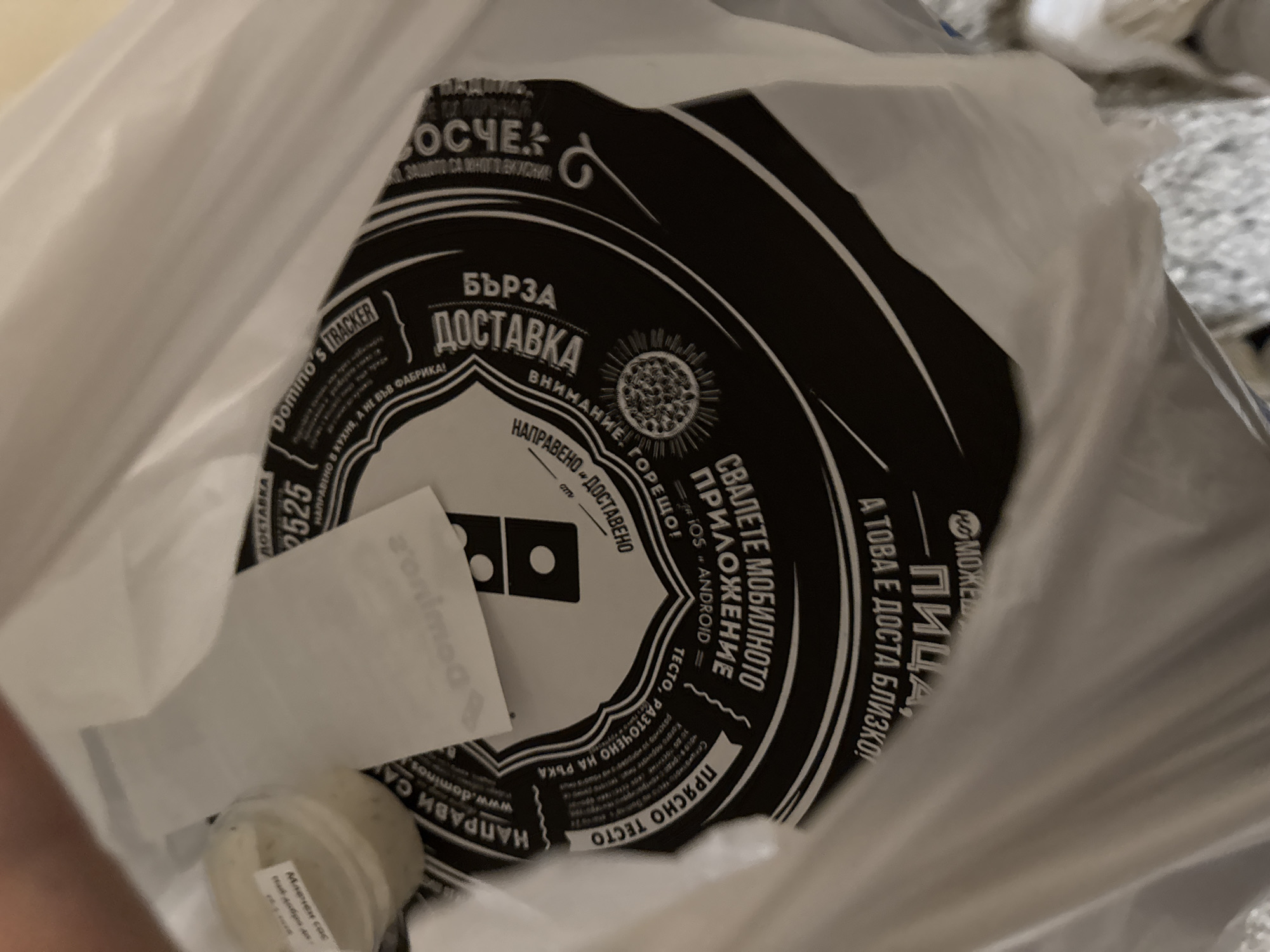 The image shows a Domino's Pizza delivery bag, likely photographed from inside. Visible inside the bag are a black and white round container lid with Domino's branding and Bulgarian text ("Бърза доставка" meaning "Fast Delivery," and "Внимание, горещо!" meaning "Attention, hot!"), a small container of "Млечен сос" (Milk Sauce) and a Domino's Pizza paper bag insert promoting their app. The Bulgarian text suggests this is likely a Domino's Pizza branch in Bulgaria, perhaps in the city of Sofia, where Domino's has locations.