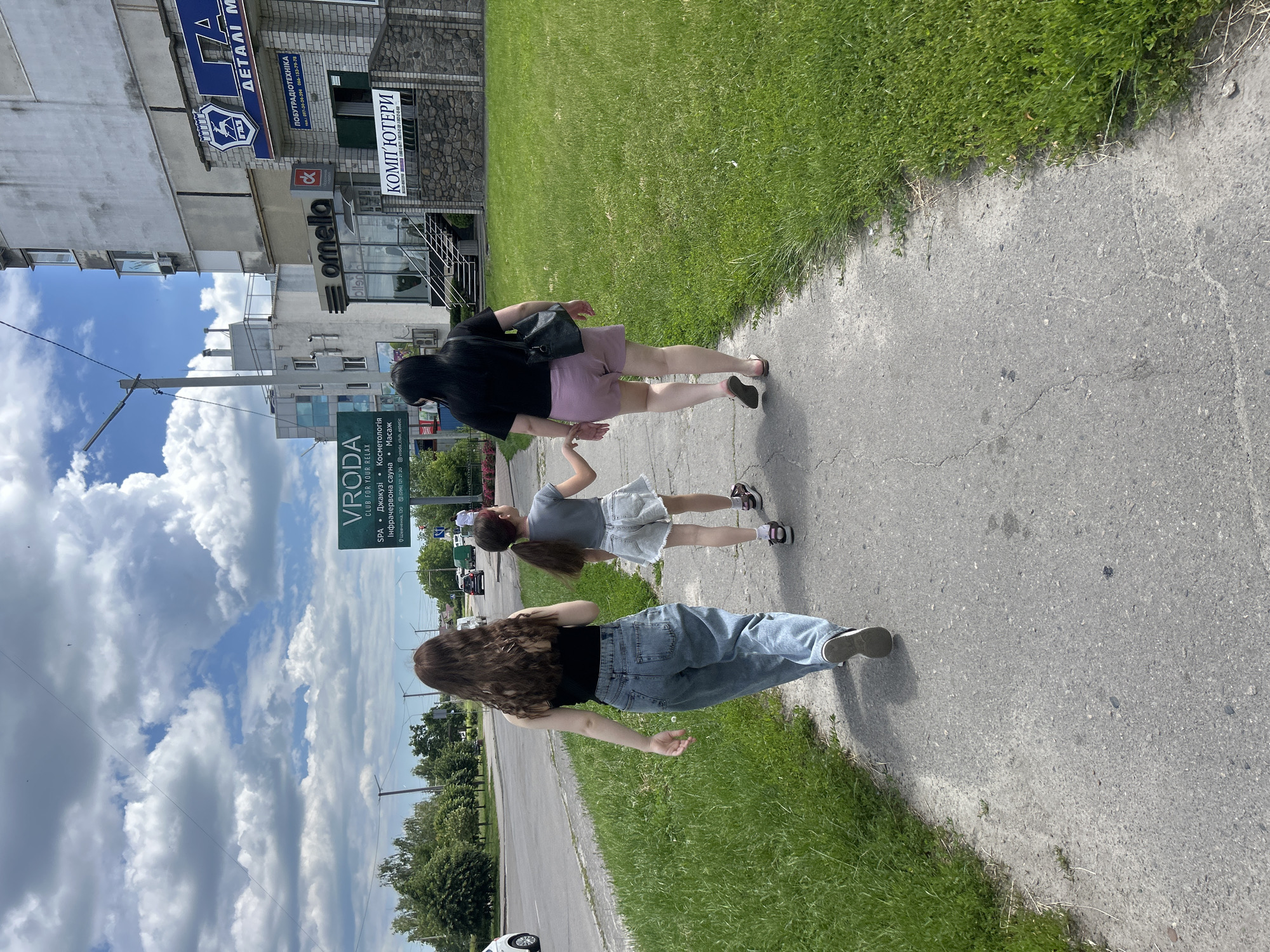 Three individuals, two women and a young girl, are walking away from the camera on a sidewalk. The sidewalk is adjacent to a grassy area and a street with light traffic. In the background, there are buildings, including one with the signage "VRODA CLUB FOR YOUR RELAX" and another that appears to be a commercial establishment with "КОМП'ЮТЕРИ" (Computers) visible. The sky is partly cloudy. The location is identified as Bila Tserkva, Ukraine. The woman on the left wears a black tank top and light blue jeans. The woman on the right wears a black t-shirt and pink shorts. The girl in the middle wears a gray t-shirt and light-colored denim shorts. She is holding hands with the woman on the right.