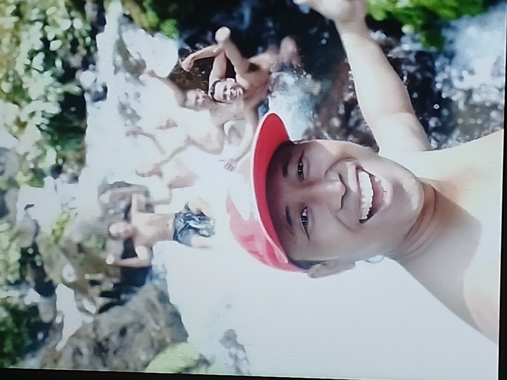 The image shows a selfie of a person in the foreground wearing a red baseball cap and smiling. In the background, several shirtless men are in a body of water, possibly a waterfall or river, with lush green foliage on the right side and rocky formations. The water appears to be flowing, creating white splashes. The men in the background are engaging with the water, with some appearing to be splashing or posing. The overall scene suggests an outdoor recreational setting. The image quality is low, with visible pixelation and a lack of sharp detail. No specific location in Surabaya, Indonesia can be identified from the visual information present.