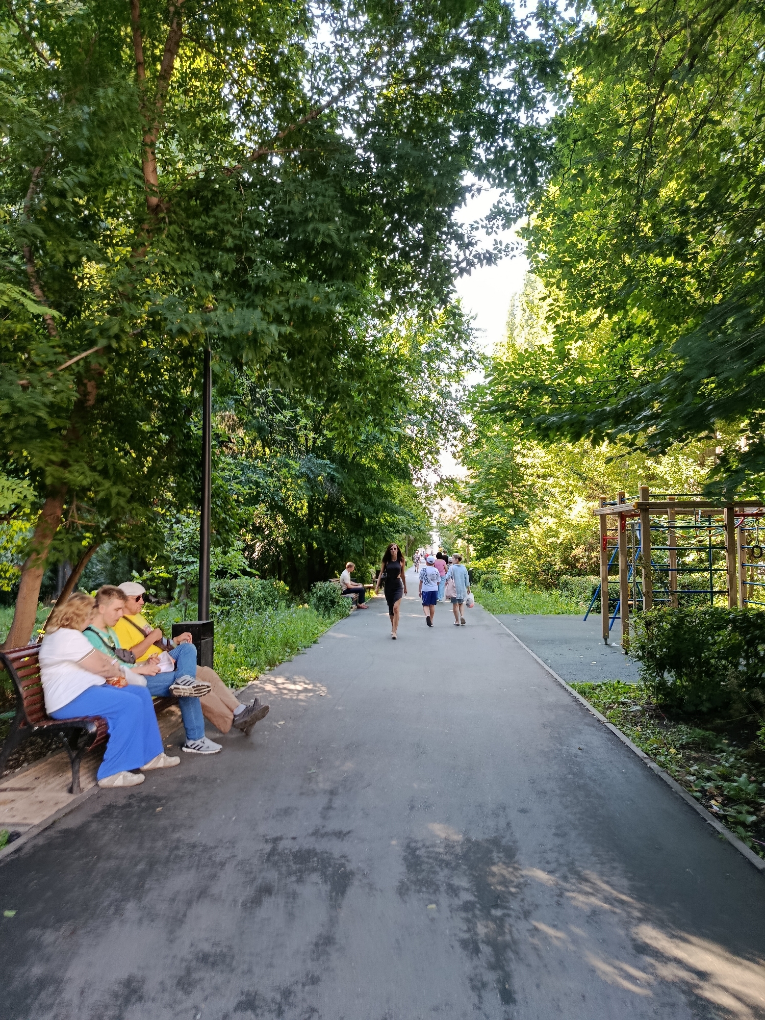 In a sun-dappled park, a paved walkway curves through lush green trees. On the left, a wooden bench seats three individuals: a woman in a white shirt and blue pants, a man in a yellow shirt and jeans, and a young person in a green shirt. The woman and young person appear to be an adult and child. The man is seated between them, with his arm around the young person. Further down the path, several people walk in the distance, including a woman in a black dress, a woman in a pink shirt and shorts, and a woman in a blue top and white shorts. To the right of the walkway, a children's playground structure with wooden beams and ropes is visible, partially obscured by dense foliage. The ground of the walkway is wet in patches, reflecting the dappled sunlight from the trees. The location is identified as the Children's Park on Volgogradskaya Street in Saratov, Russia.