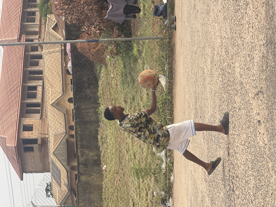 A young male is depicted mid-stride on a coarse, light-colored asphalt surface in Umunekwu, Nigeria. He wears a patterned short-sleeved collared shirt, white shorts with blue trim, and dark grey slip-on shoes. His right hand holds a worn orange basketball extended forward and slightly downwards, with his left foot positioned ahead, suggesting an impending dribble or pass. Behind the individual, a chain-link fence separates the foreground from a grassy area containing sparse vegetation and scattered white plastic litter. In the background, beyond the fence, a lower single-story building with multiple yellow-tiled gabled roofs and rounded arch windows is visible, with solar panels installed on one roof section. Above and behind this, an unfinished two-story concrete block structure with a red-tiled roof and open window frames stands. To the far right, a pole is present, and a grey shirt and dark pants hang from an unseen support, with a dark backpack and sandals resting on the ground nearby. A reddish-brown dirt mound is situated behind the pole.