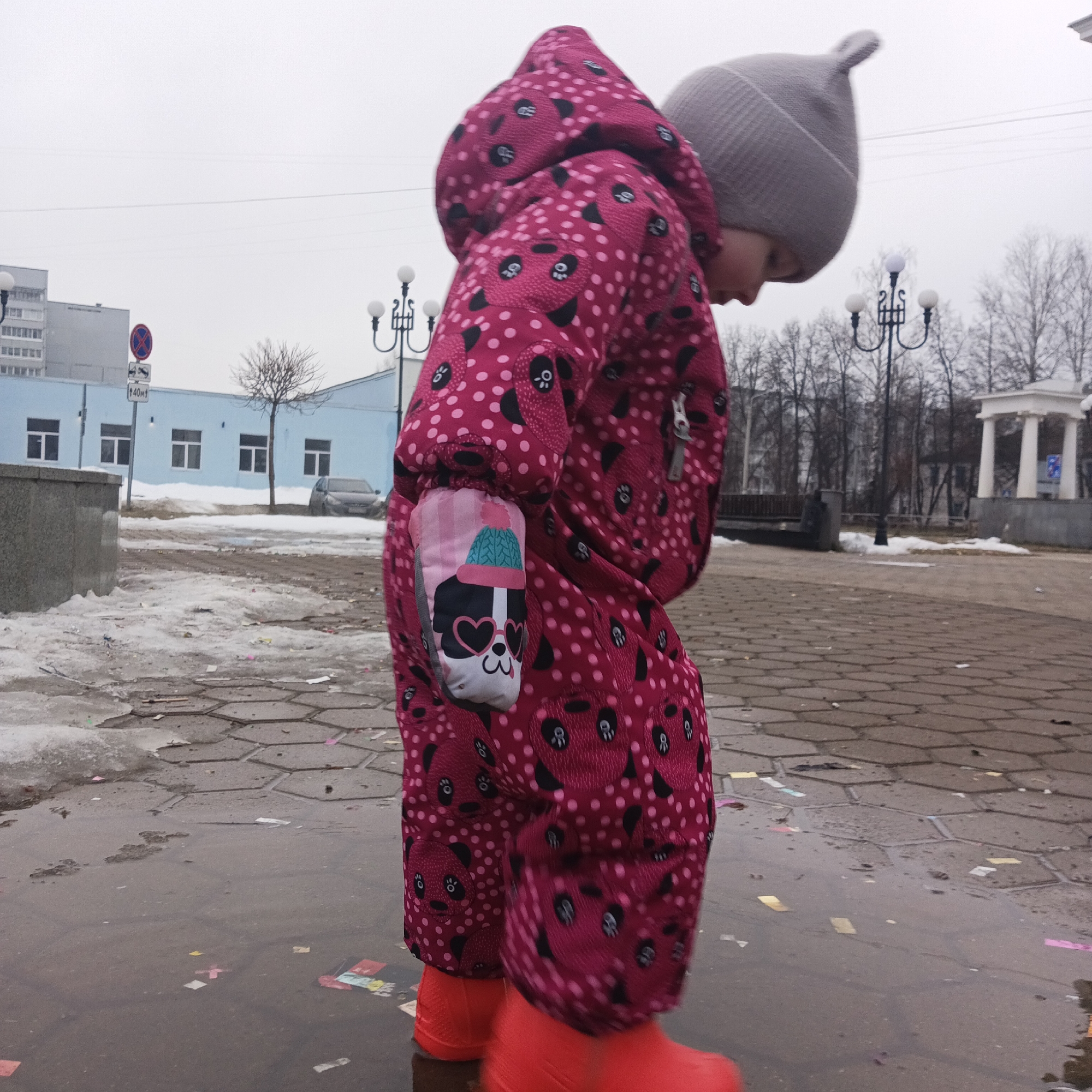 A small child wearing a pink snowsuit with a panda design, a grey knit hat, and bright red rubber boots is standing in a puddle in Chaikovsky, Russia. The puddle is on a brick-paved area strewn with confetti. In the background, a light blue building, street lights, bare trees, and a white rotunda structure are visible. The sky is overcast. The photo appears to be taken at ground level.