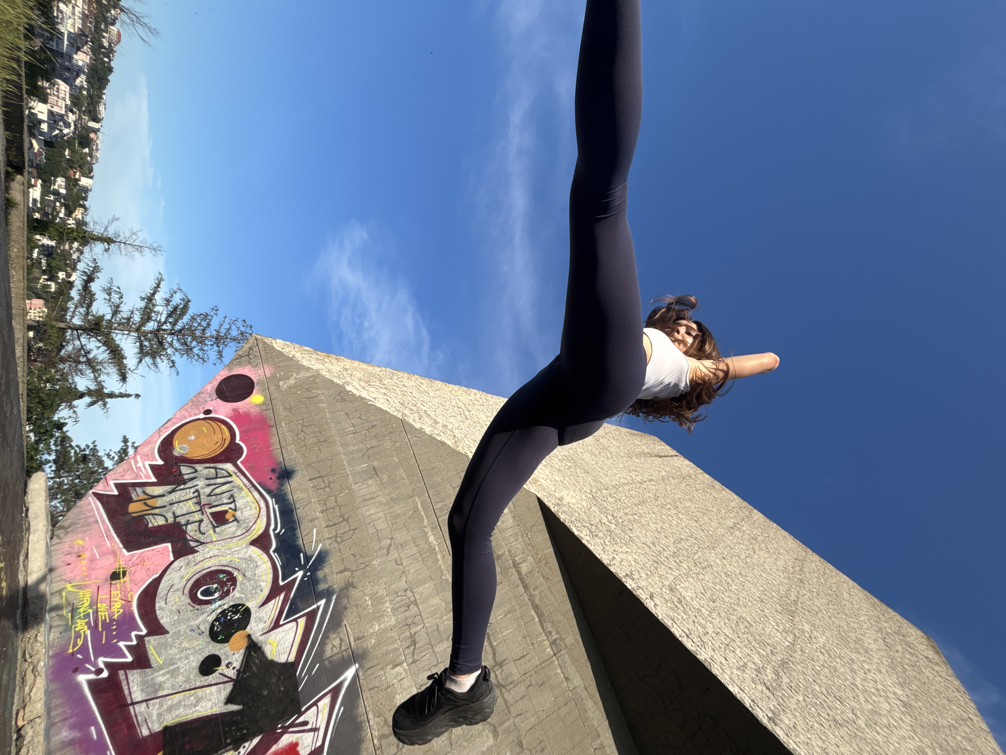 A young woman with brown hair performs a split jump in mid-air against a clear blue sky. She is wearing a white tank top and dark leggings, with black athletic shoes. The background features a Brutalist concrete structure adorned with graffiti, and a cityscape of Varna, Bulgaria, with buildings on a hillside. The concrete structure's rough texture and geometric design are visible, as is the colorful graffiti on its lower section. The lower portion of the image shows trees and a grassy area.