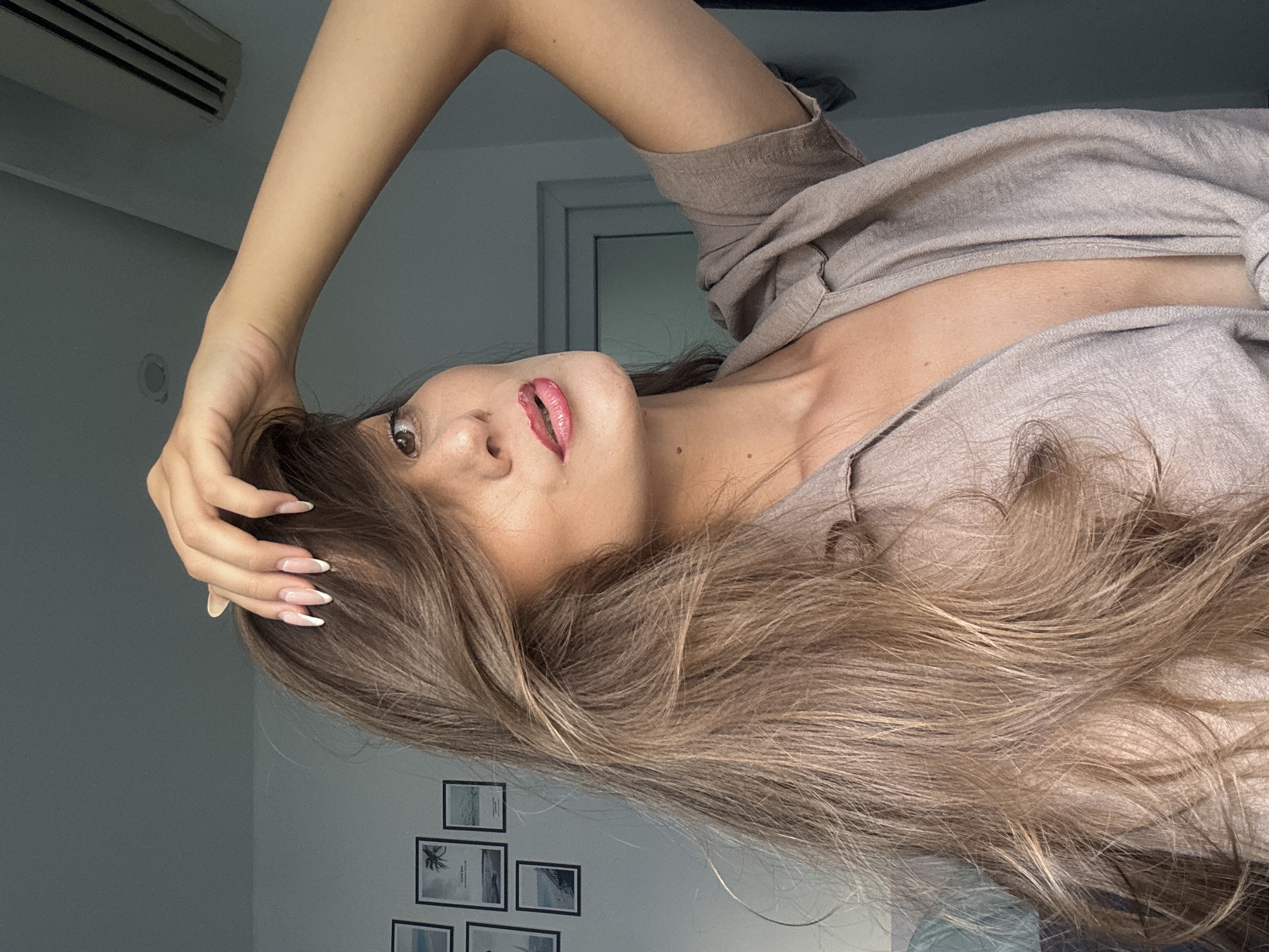 A woman with long brown hair is seen in a close-up, slightly tilted composition. She wears a taupe-colored shirt and has a hand near her head. Her nails are manicured with a French tip design. The background consists of a wall decorated with a grid of small framed pictures. In the upper left corner, an air conditioning unit is visible. The location cannot be determined from the visual information.