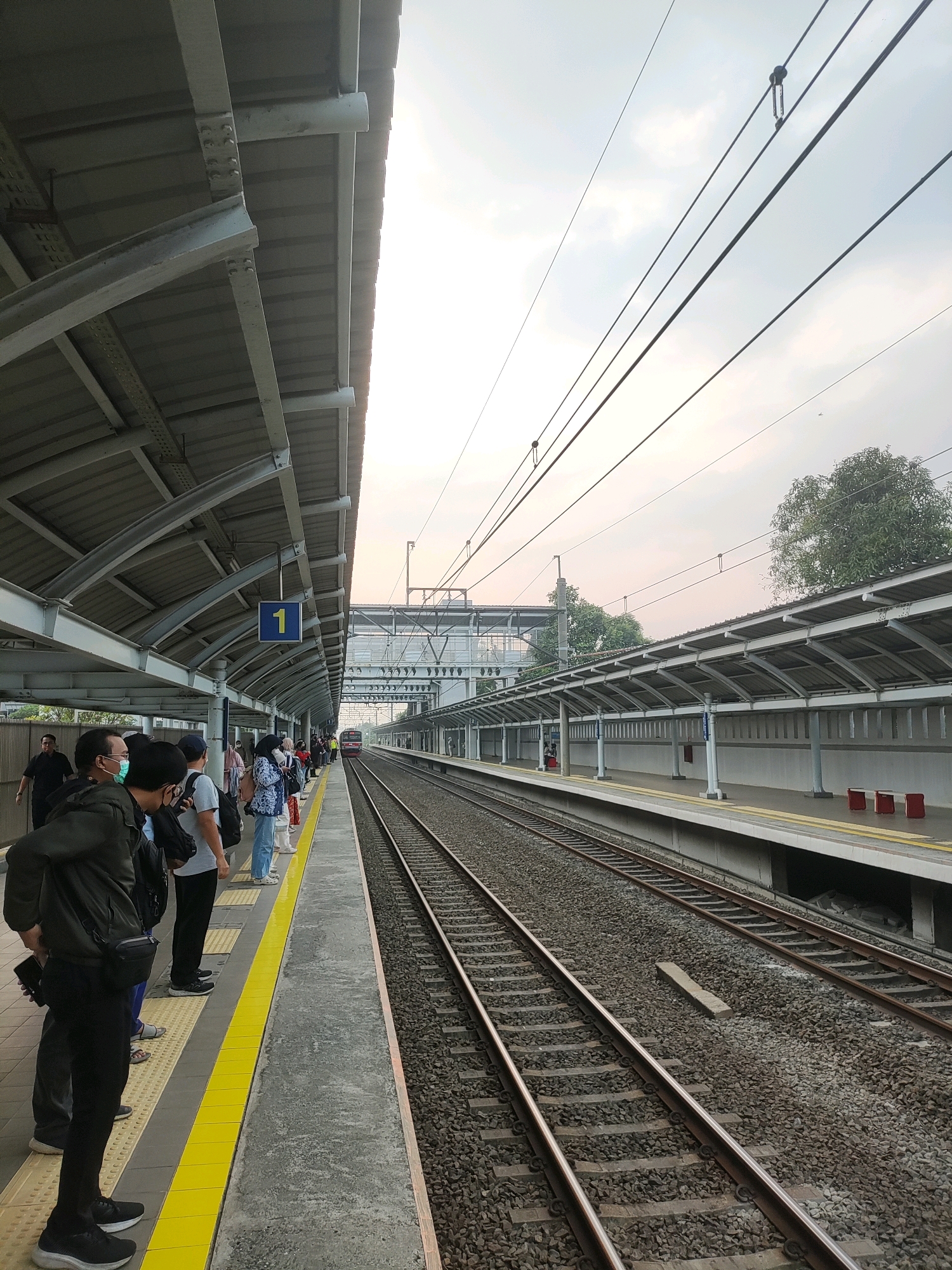 This is a photograph of a train station platform in Telagamurni, Indonesia, during daylight hours. The platform is covered by a gray metal roof supported by a steel frame, with electrical wires visible above. Several people are standing on the platform, waiting for a train, some wearing face masks. The platform edge is marked with a yellow line. Adjacent to the platform are train tracks laid on a ballast bed, with a red train visible in the distance. The sky is overcast with a pale orange hue, suggesting either dawn or dusk. Trees and foliage are visible in the background.
