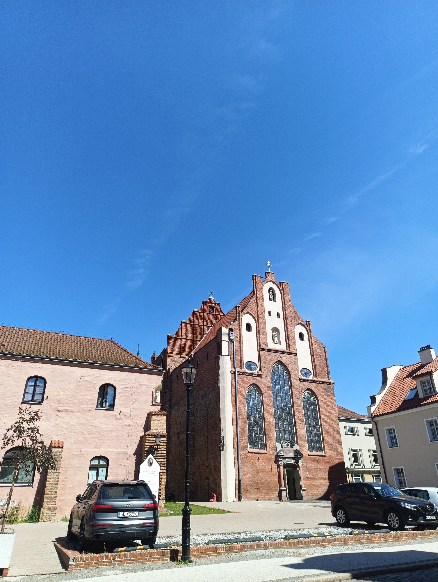 This image captures an outdoor urban scene on a clear, sunny day. The focal point is a large, red brick Gothic church with a stepped gable and a prominent cross at its apex, situated in Gdańsk, Poland. Adjacent to the church is a lighter-colored, three-story building with arched windows. In the foreground, a dark gray Audi SUV is parked. Another dark SUV, possibly a Mazda, is parked further to the right, alongside a white van. A black lamppost stands between the buildings and the vehicles. The ground is paved with brick and stone. The sky is a vibrant, cloudless blue.