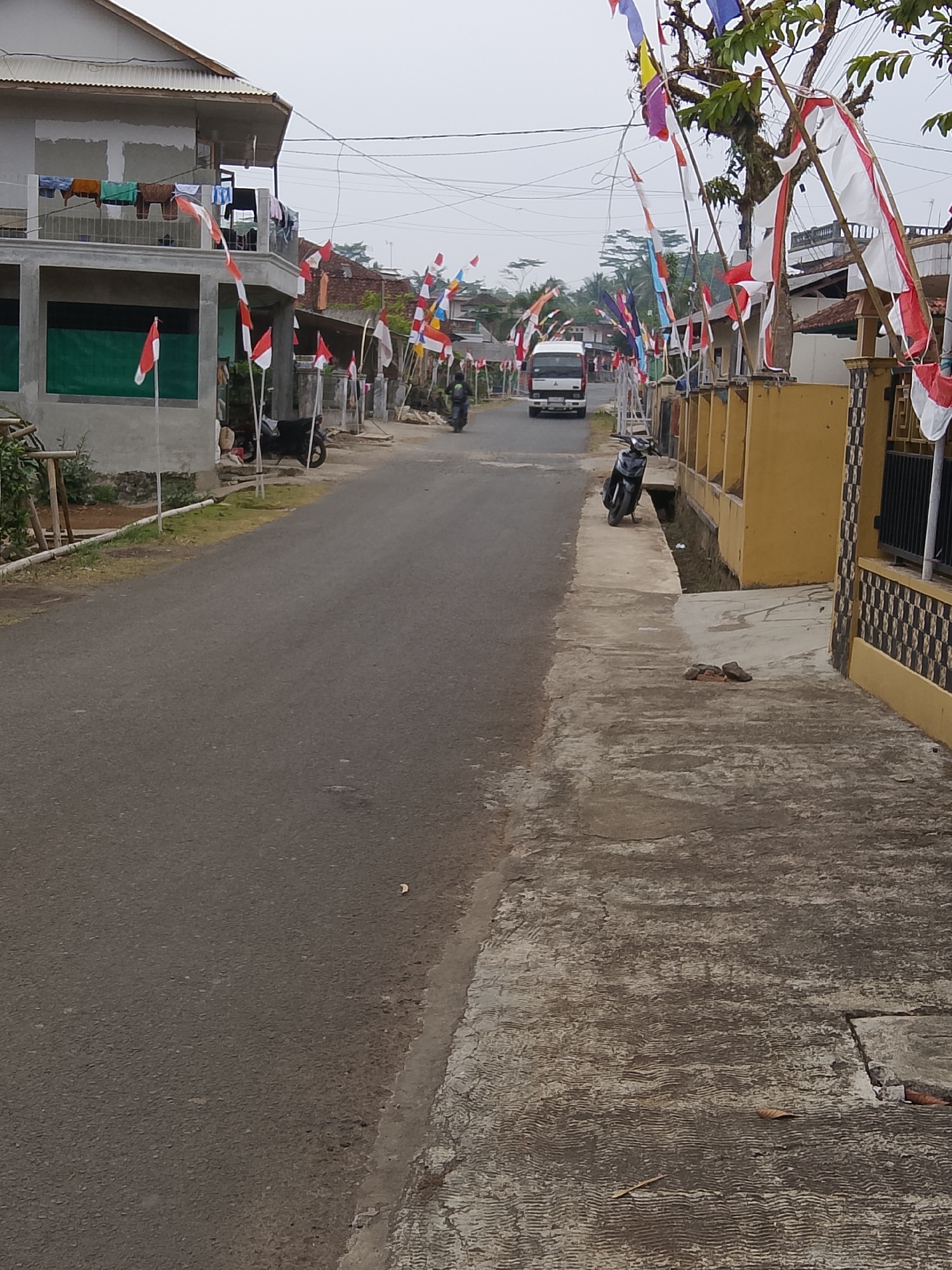 In the city of Cikukulu, Indonesia, a street is lined with Indonesian flags and festive decorations. The scene captures a residential area, possibly during a national celebration. A two-story house is visible on the left, with laundry hanging on a balcony. On the right, a yellow building with a checkered pattern borders the street. Several motorcycles are parked along the roadside. A white truck is driving down the street in the distance. The sky is overcast, suggesting a cloudy day. The ground is paved with asphalt and concrete.