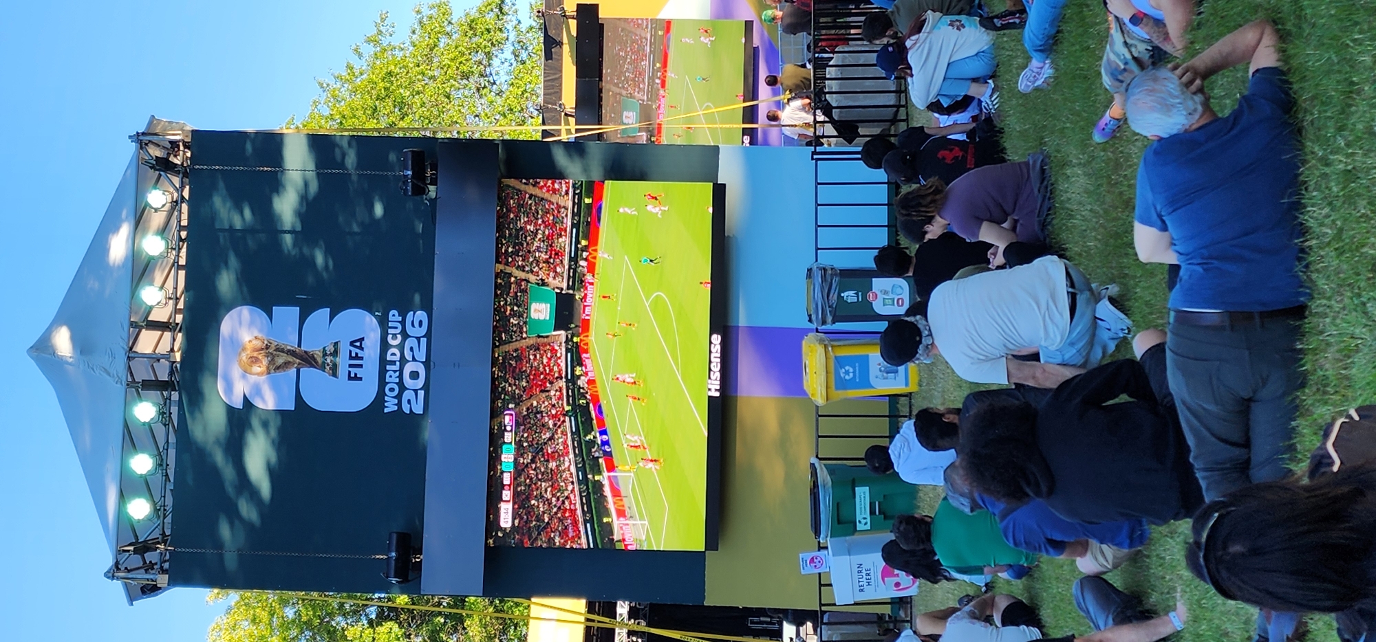 An outdoor public viewing area features a large screen displaying the FIFA World Cup 2026 logo and a soccer match. Spectators are seated on the grass in front of the screen, facing it. The screen shows a soccer game in progress with players on the field and a stadium filled with fans in the background. The FIFA World Cup 2026 logo is prominent above the game footage. Behind the spectators, there are several trash and recycling bins, and a metal fence. The area appears to be a park or an open public space, with trees visible in the background and a clear blue sky.