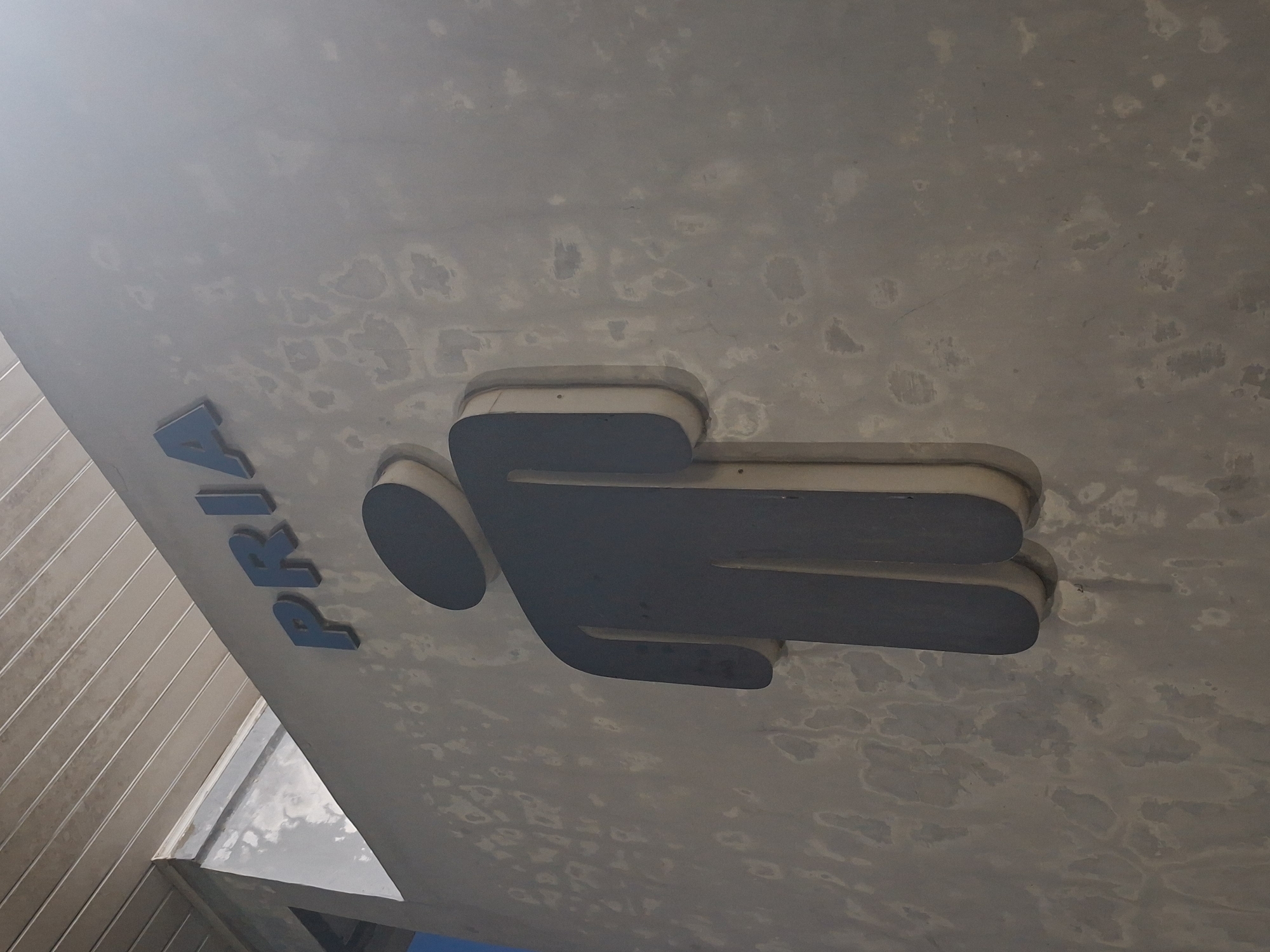 A sign indicates a men's restroom at a facility in Sinyeu, Indonesia. The sign features a dark grey silhouette of a person and the blue word "PRIA," which means "men" in Indonesian. The sign is mounted on a rough, light-colored wall that shows signs of wear and discoloration. Above the wall, a portion of a light-colored ceiling with horizontal slats is visible. The overall environment appears to be outdoors or in a covered outdoor area, given the lighting and the visible ceiling structure.