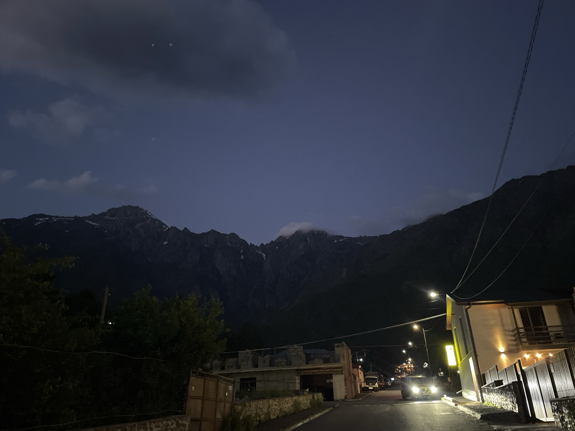 A nighttime view of a street in Stepantsminda, Georgia, faces a dramatic mountain range. The sky is a deep twilight blue with scattered clouds and visible celestial bodies, including a crescent moon and a few faint stars. The street is paved and curves slightly to the right. On the right side of the street, a modern building with illuminated windows and balconies stands out. A fence made of metal and stone borders the property. Streetlights cast pools of yellow light onto the road. Several vehicles are present, including a car with its headlights on driving down the street, and two trucks in the distance. On the left side of the street, a partially constructed building made of concrete blocks is visible, along with trees and a fence. The imposing, dark mountain range, partially covered in snow at its peaks, dominates the background.