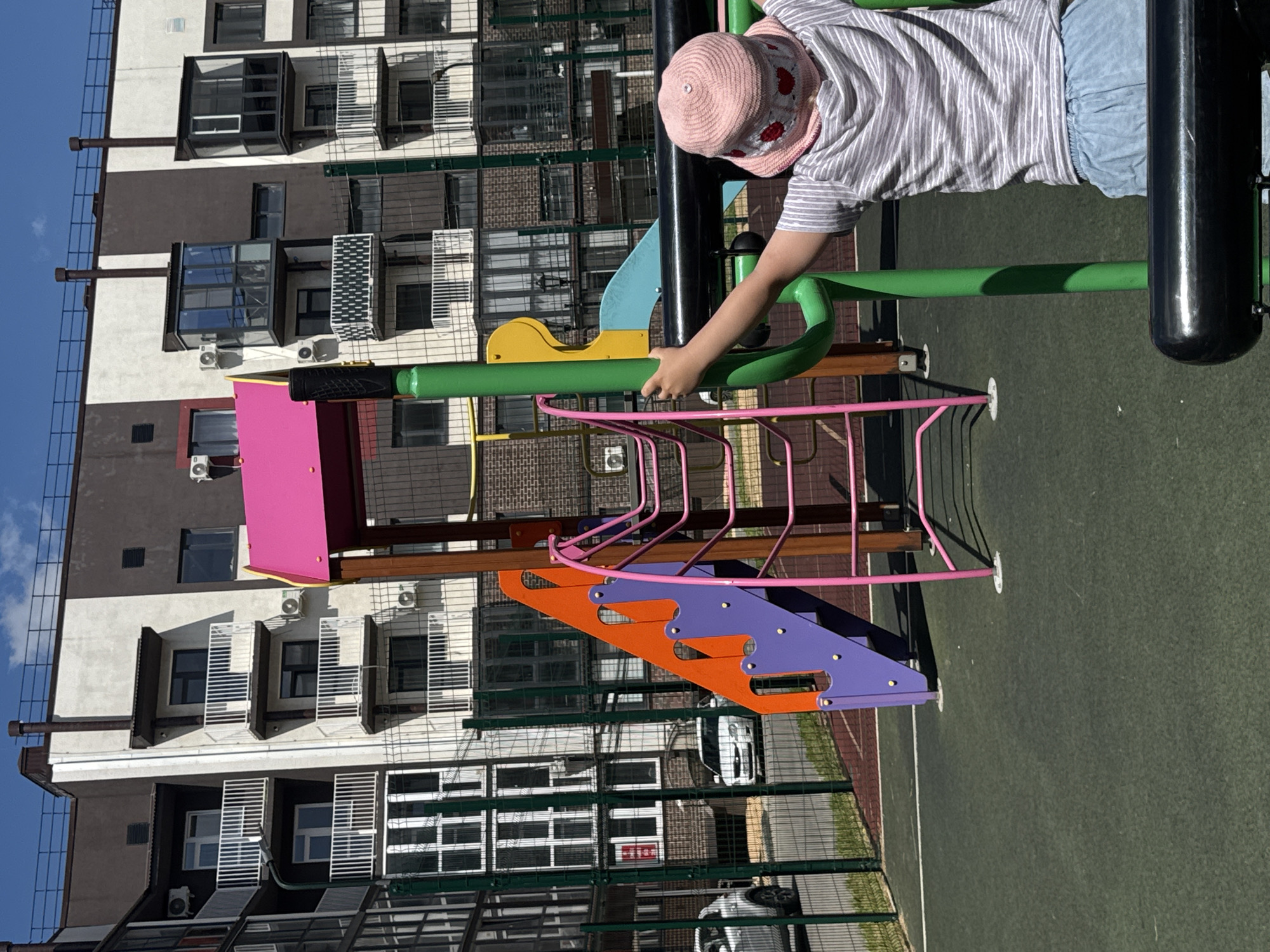 The image shows a child in a pink hat and a striped shirt playing on a colorful playground. The playground has bright pink and purple plastic steps, orange accents, and green metal bars. The child's hand is gripping a green metal bar. In the background, there is a modern apartment building with balconies and air conditioning units. The sky is blue and appears clear, suggesting a sunny day. The playground is situated on a green, soft surface, likely for safety. The location is identified as Вязники, Russia. The image is taken from a slightly elevated, angled perspective, capturing both the child and the surrounding environment.