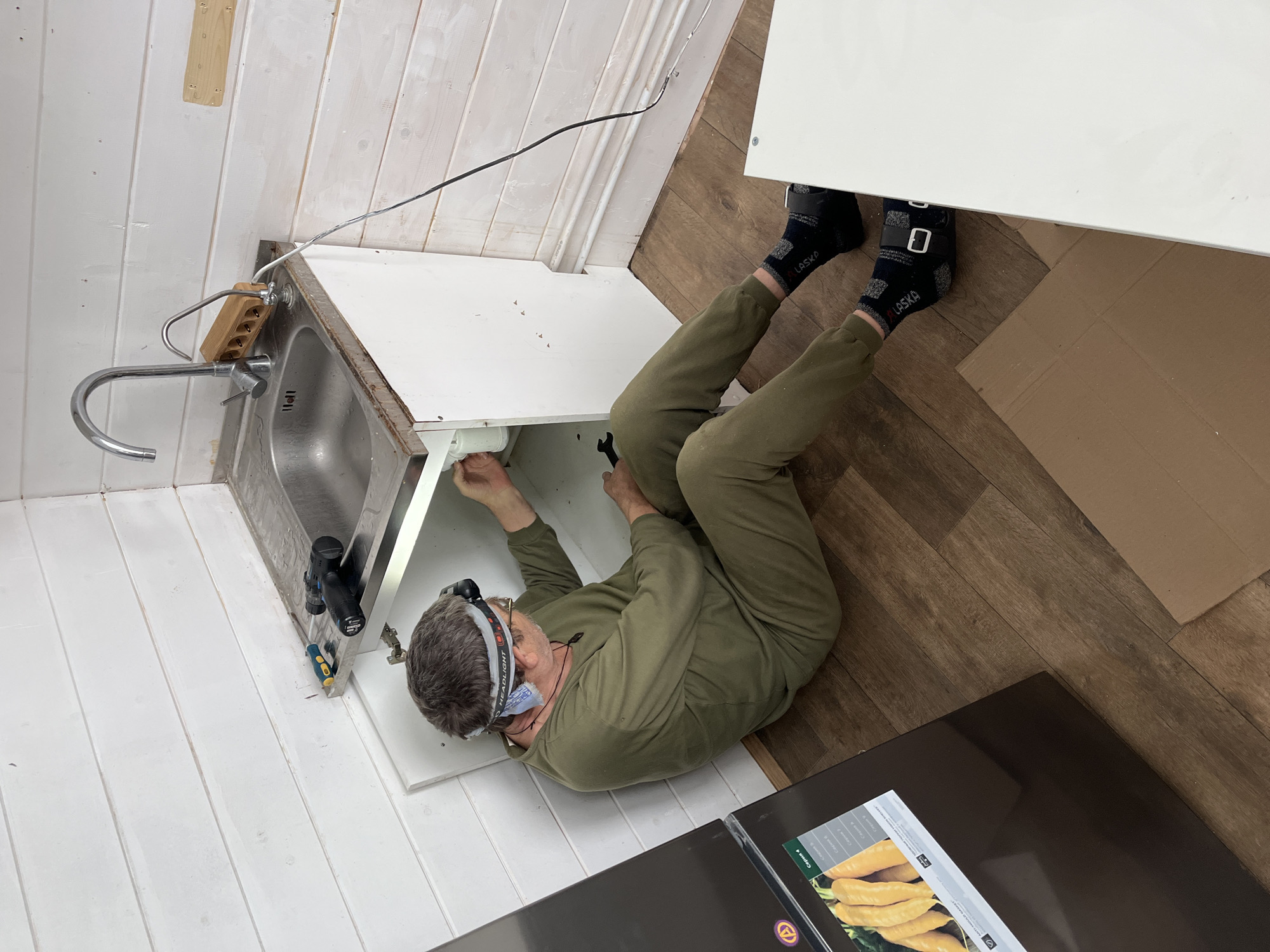 A middle-aged man wearing green overalls and black socks is lying on his back on a wood-look floor. He is under a white kitchen sink cabinet, using a wrench to work on the plumbing. The sink is stainless steel and the wall is white with horizontal planks. Tools are visible near the sink. The location is Раздолье, Russia.