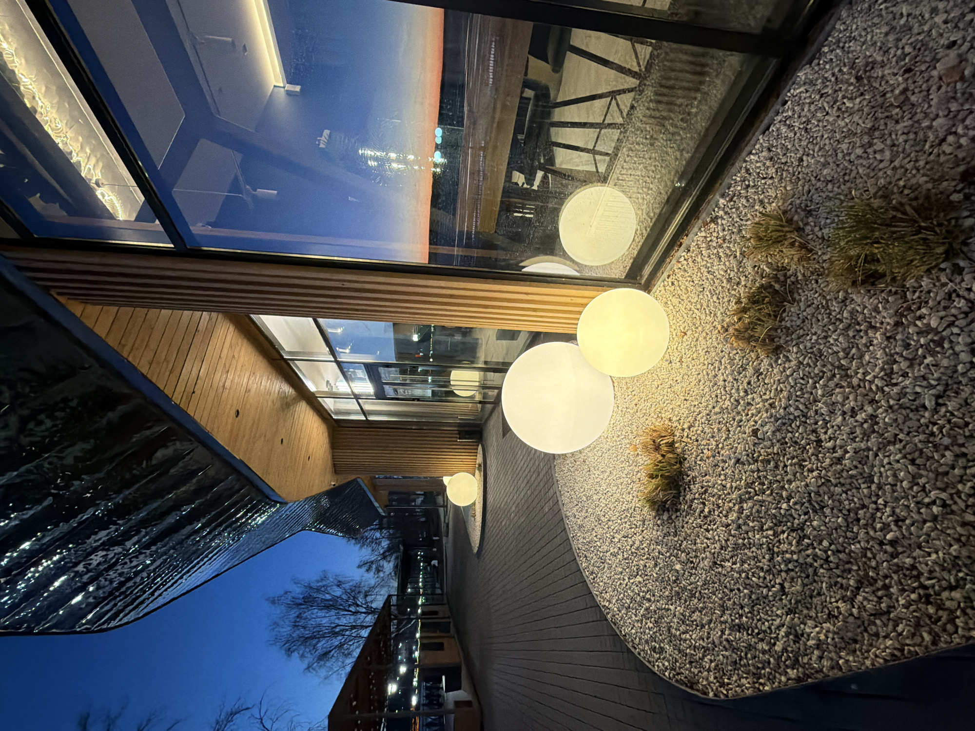 The image captures an architectural scene, possibly at twilight or in the evening, featuring a modern building design with a combination of glass, wood, and dark paneling. A notable feature is the large window reflecting a cityscape with illuminated buildings against a darkening sky. Adjacent to the building, an area covered in small stones contains spherical lights, with a few patches of greenery interspersed. The scene suggests a contemporary design aesthetic, combining natural elements with modern architectural forms. Based on the prompt, this location may be in Votkinsk, Russia, though no distinct landmarks from the image allow for its confirmation.