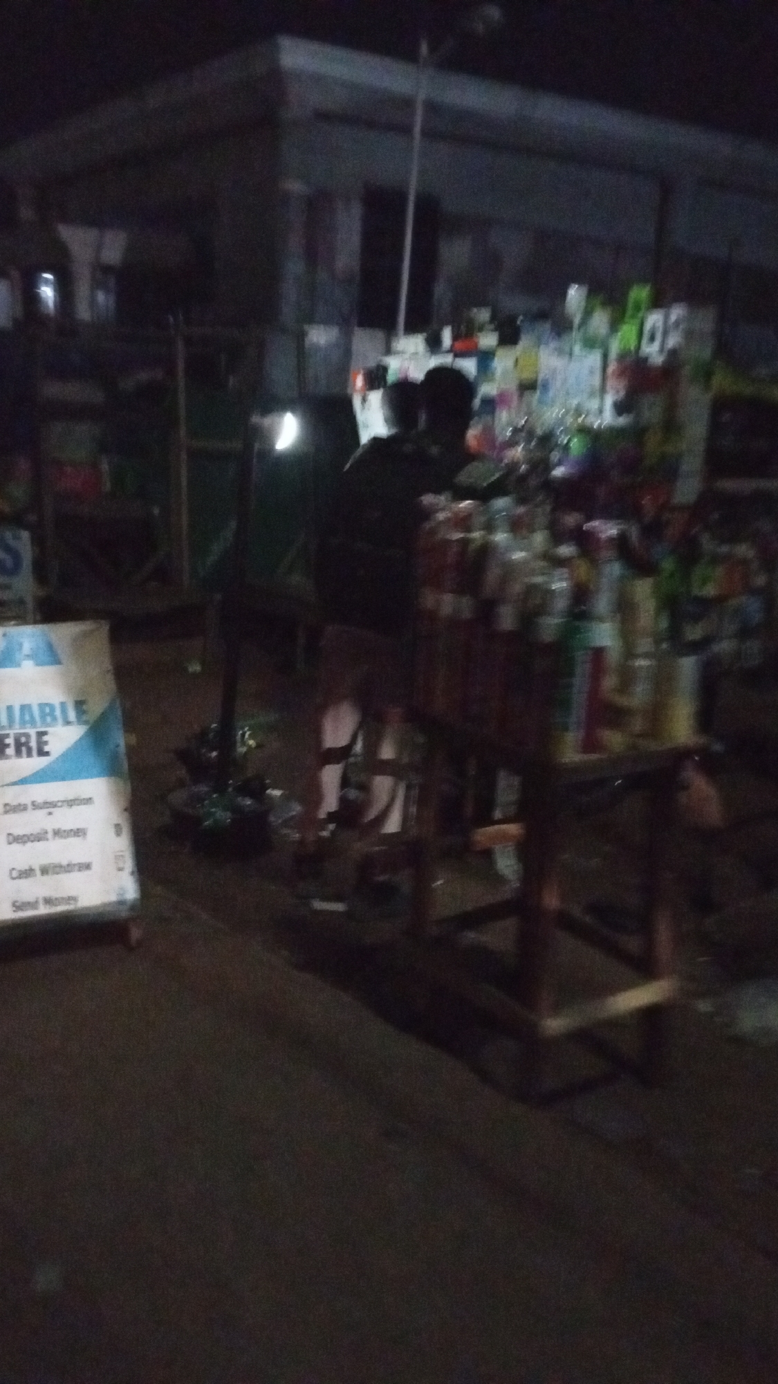 This dark and blurry image captures a street scene at night, likely in Lokoja, Nigeria. The primary focus is a vendor's stall and a nearby sign, both dimly lit.
In the foreground on the left, a white and blue sign is partially visible, indicating a mobile money or agency banking service. The legible text on the sign includes: "LIABLE HERE", "Data Subscription", "Deposit Money", "Cash Withdraw", and "Send Money". This suggests the presence of a point for financial transactions or mobile services.
To the right of the sign, a person, seen from behind, appears to be attending a wooden stall. This stall is densely packed with numerous small bottles, possibly containing perfumes, oils, or other small retail items, arranged on multiple shelves. The stall and the person are illuminated by an unseen light source, casting soft glows and shadows.
In the upper-left portion of the background, a distinct, bright, crescent-shaped light source is visible, which could be a moon or a powerful street lamp. The overall background consists of dark, shadowy shapes, likely other buildings or market structures, further emphasizing the nighttime setting. The image's blurriness and low light conditions convey the atmosphere of an active, albeit dimly lit, evening street or market.