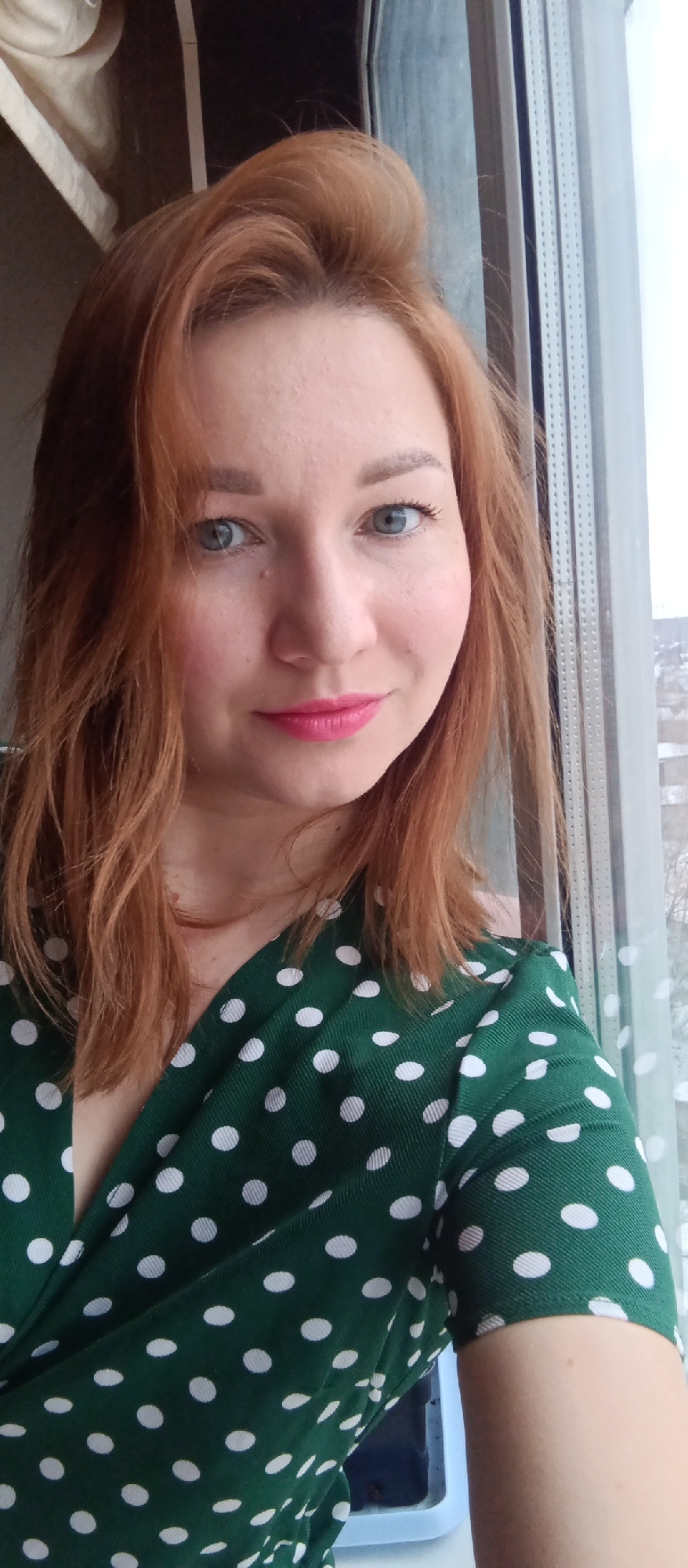 A selfie of a woman with reddish-brown hair, blue eyes, and pink lipstick, wearing a green dress with white polka dots. The woman is positioned in front of a window. Through the window, a cityscape is visible.