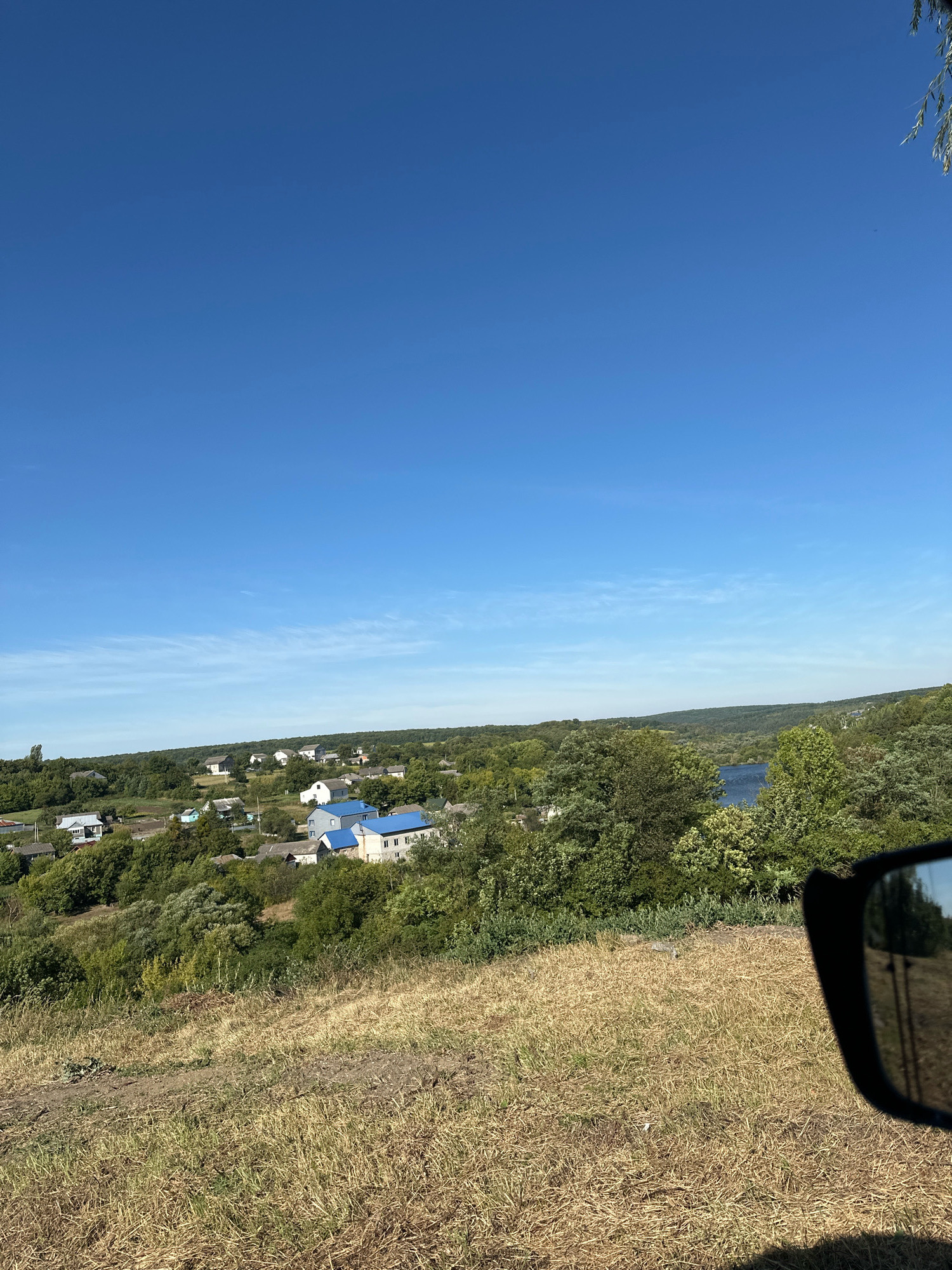 The image depicts a rural landscape with scattered houses and a body of water, possibly a river or lake, in the distance. The scene is viewed from a slightly elevated perspective, with a grassy slope in the foreground. The sky is clear and blue with some wispy clouds. In the middle ground, a cluster of houses with blue and grey roofs are visible amidst lush green trees. A car's side mirror is visible in the lower right corner, reflecting trees and a dirt road. The location is identified as Makiv, Ukraine. The overall impression is one of a peaceful, sunlit day in a rural setting.