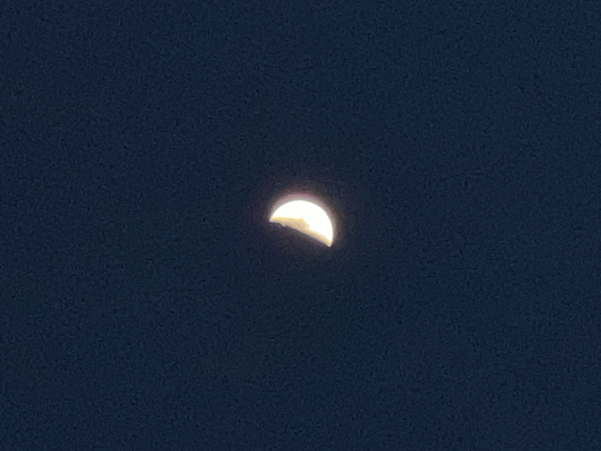 A crescent moon is visible in a dark blue sky. The moon is illuminated on its right side, with the left side in shadow. The sky appears to be clear, with no other celestial bodies visible. The image is likely taken at night, possibly in a rural or remote location given the lack of light pollution.