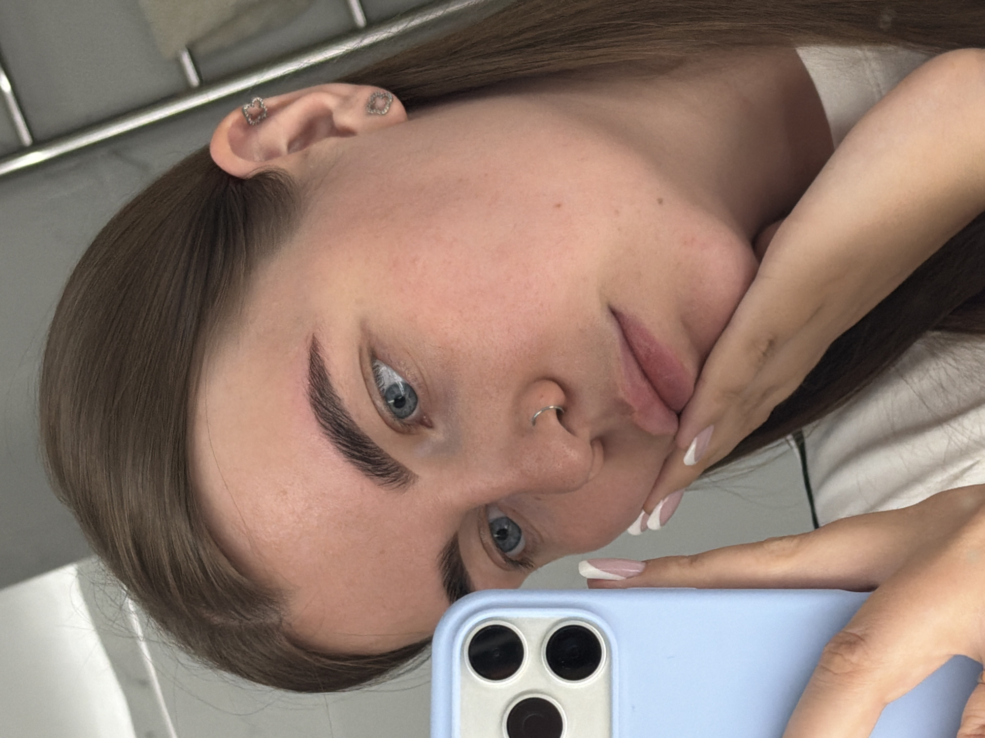 A young woman is taking a selfie with a blue smartphone in what appears to be an indoor setting, likely a room. She is fair-skinned with light brown hair, blue eyes, and distinct eyebrows. A nose ring is visible on her left nostril. She has multiple ear piercings with small heart-shaped earrings. Her fingernails are manicured with a French tip design. She is wearing a white top. While the setting provides little to identify a specific location, the prompt requests the location as Chaikovsky, Russia.