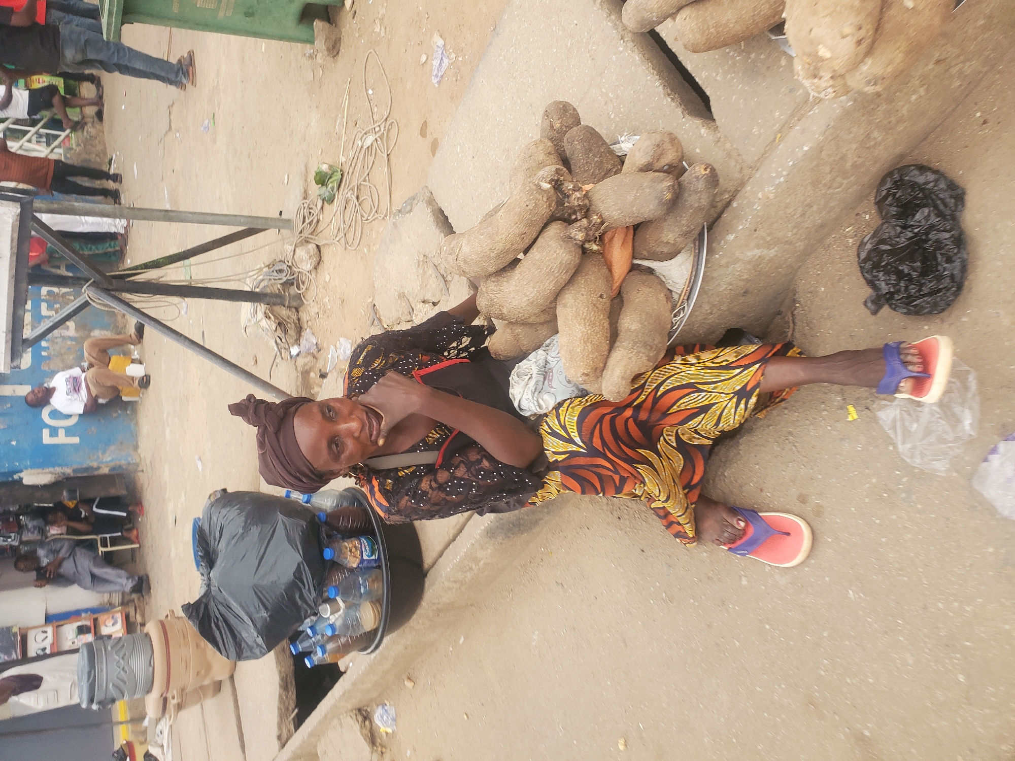 This candid, rotated image captures an outdoor market scene, likely in Jos, Nigeria, during daylight hours under clear weather conditions. The central subject is a woman, appearing to be a vendor, sitting on the ground and looking directly at the camera with a neutral to slightly smiling expression. She is dressed in a vibrant patterned skirt, a dark long-sleeved top with white polka dots, and a brown headwrap. She wears red and blue flip-flops.
Beside her, to her right, is a large metal tray heaped with numerous yams, indicating her primary merchandise. To her left, another tray (or part of one) holds several plastic bottles, some containing clear liquid (possibly water) and others with a darker substance, along with a black plastic bag.
The setting is a dusty, unpaved area beside a concrete curb or raised path. In the background, various other people are visible, some standing, some seated, suggesting a bustling market environment. Metal poles, possibly part of temporary stalls or structures, crisscross the middle ground. Tangled ropes and some debris are scattered on the ground. Part of a green trash can is also visible. Further back, a building or shop with a blue facade shows the partial text "FO," likely part of a larger word. The overall atmosphere suggests a typical informal urban street market.