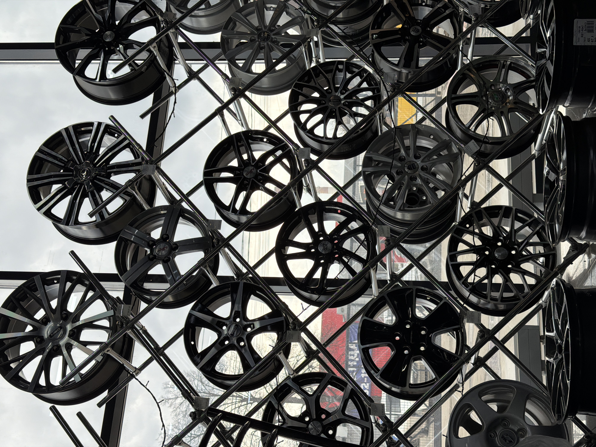 The photograph depicts a display of car wheels hanging on a metal rack, likely within a store or showroom. The wheels are mostly black and feature various spoke designs. The background is a window looking out onto a city street, partially obscured by the displays. The presence of text in Russian, along with visible urban architecture and signage, suggests the image was taken in a city. Considering the location specified, the image is most likely to be taken in Izhevsk, Russia. The wheels are suspended by hooks or fasteners from the metal rack, which is constructed from horizontal and diagonal bars. The street outside the window suggests an urban environment. The interior lighting is bright, creating reflections on the wheels and metal rack.