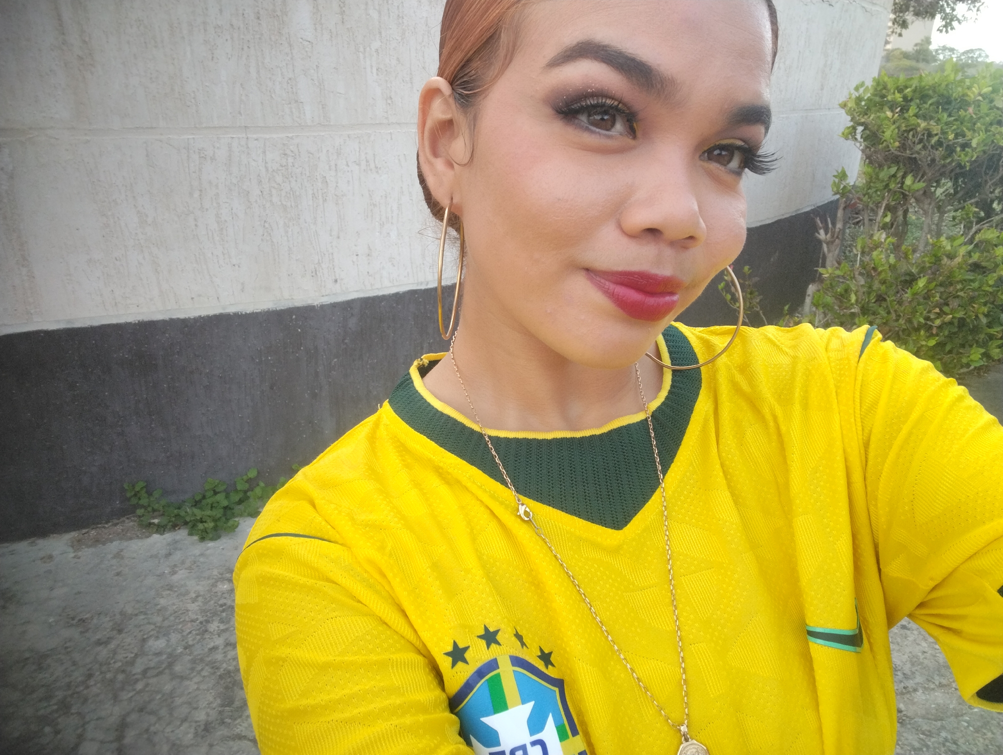 A close-up selfie shows a young woman with light brown hair pulled back and wearing bold makeup, including dramatic eyelashes and red lipstick. She has large gold hoop earrings and a gold chain necklace. She is wearing a bright yellow t-shirt with a dark green collar and trim, featuring a crest with stars and the word "BRASIL" on the left side of her chest, suggesting she is wearing a Brazilian national team jersey. The background consists of a textured light-colored wall and a dark gray textured surface below it. To the right, green foliage from a bush is visible. The scene appears to be outdoors, possibly in a residential area or a park in Porlamar, Venezuela, given the sunny lighting and the style of the wall.