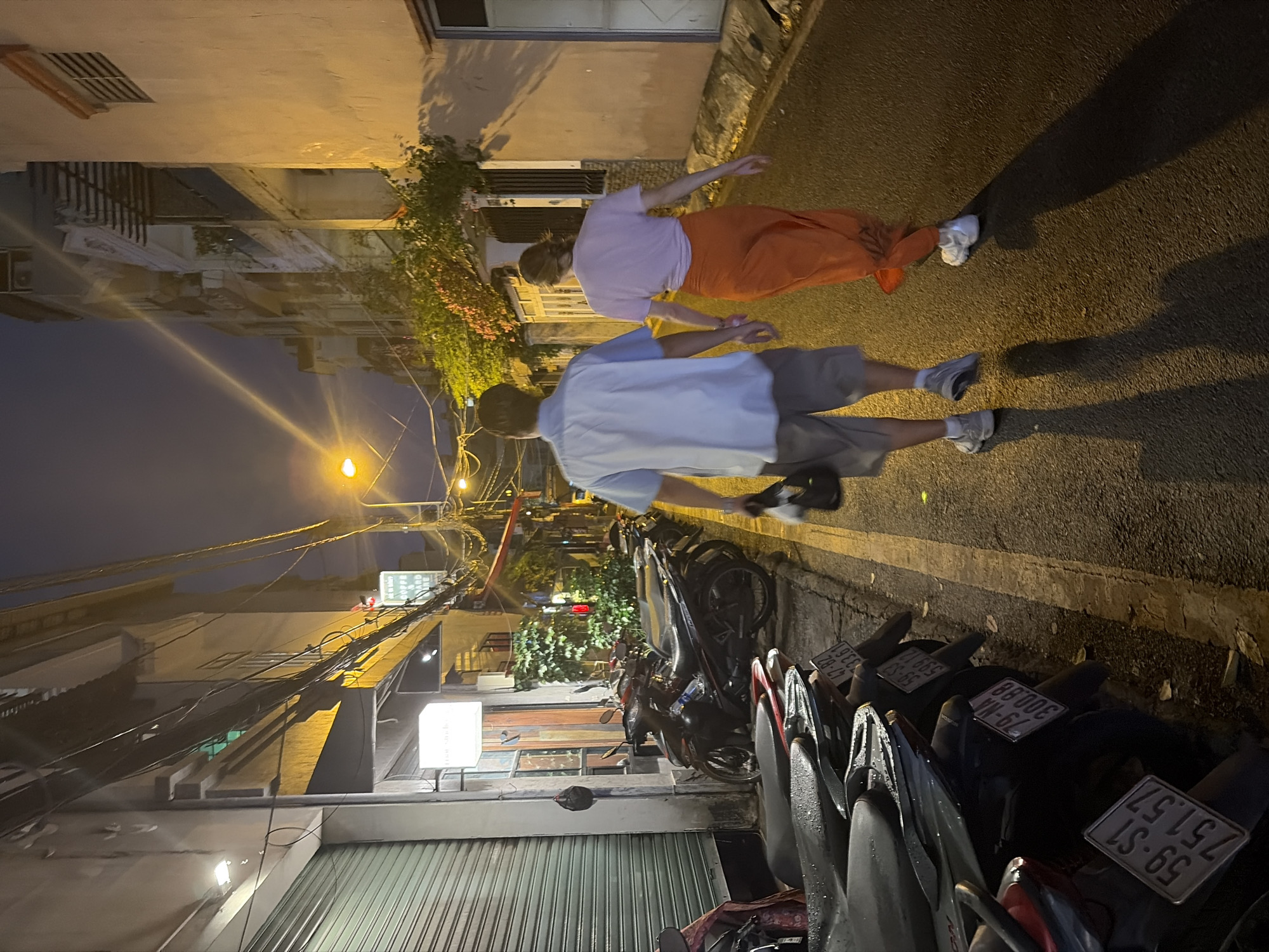 This nighttime image depicts an urban street scene in Thủ Đức City, Vietnam. Two individuals are walking away from the camera, holding hands, down a narrow asphalt road. The male on the left wears a light blue shirt, light shorts, and white sneakers, carrying a dark bag. The female on the right wears a light pink t-shirt, a long orange skirt, and white sneakers. Their elongated shadows are cast forward on the wet pavement.
To the left, a row of several motorbikes is parked on a sidewalk or shoulder adjacent to a yellow road line. Visible license plates include "59-S1 751.57", "79-MA 300.88", "59-S3 599.12", and "47-B2 333.61", with "59" indicating Ho Chi Minh City (of which Thủ Đức is a part). Behind the motorbikes, a building features a closed metal roll-down shutter and a lit square sign displaying "Năm Mười". Further along, another partially visible sign reads "COFFEE AND". Numerous tangled electrical and communication wires are strung overhead between poles and buildings.
To the right of the walking individuals, a light-colored building wall fronts the street, adorned with overgrown foliage and some plants exhibiting reddish-orange flowers. The street appears wet, reflecting the light from multiple streetlights and building illumination sources, creating strong contrasts. The sky above is dark, indicating a clear night.