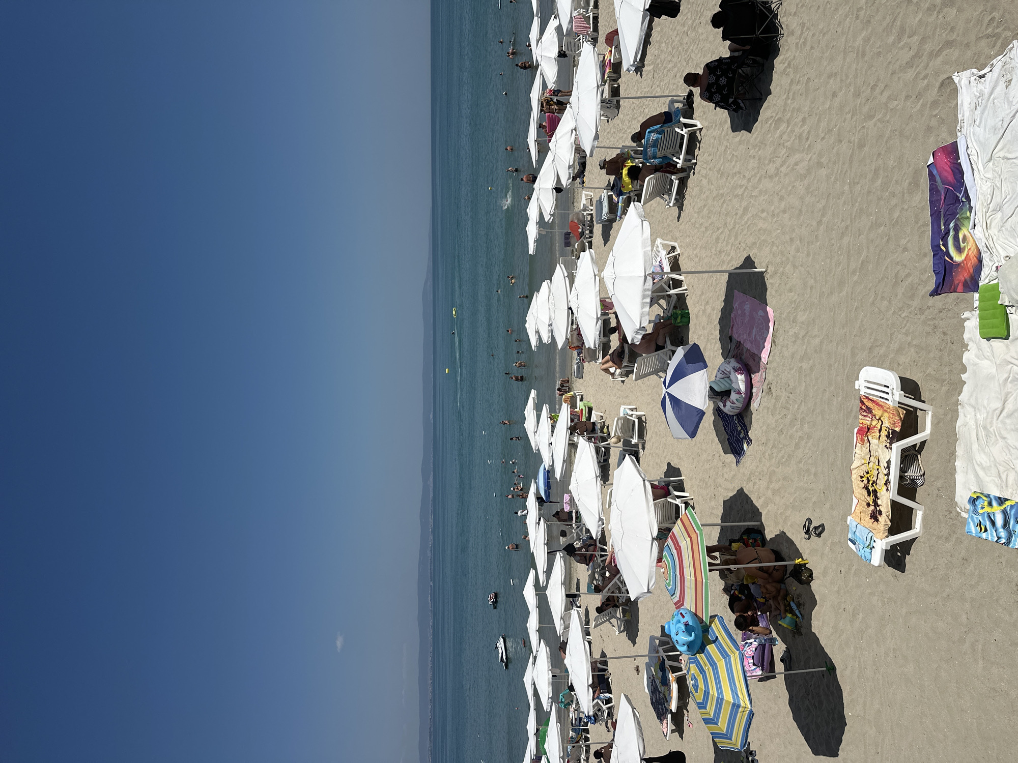 The image depicts a crowded beach scene in Pomorie, Bulgaria, on a clear, sunny day. The sand is a light beige, and numerous white beach umbrellas provide shade for rows of lounge chairs. Many people are visible, some sitting on chairs, others walking or wading in the shallow, turquoise water. Several individuals are swimming in the sea, and a few boats, including a jet ski, are present on the water. The background shows a distant coastline with low hills. Various beach accessories, such as towels, inflatable toys, and bags, are scattered across the sand. The overall atmosphere is one of a typical summer vacation day at the beach.