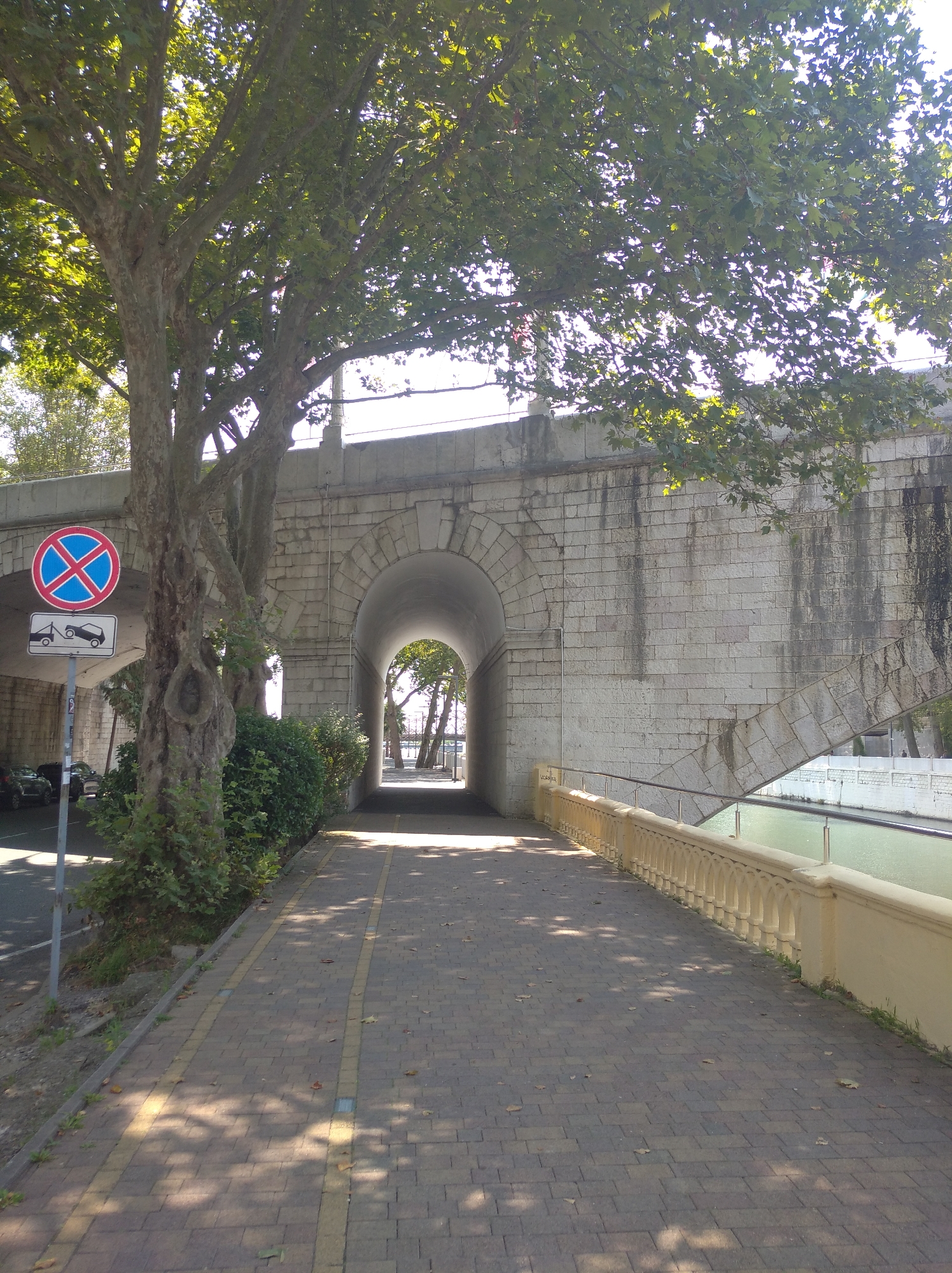 This image depicts a pedestrian walkway leading under a stone archway, likely part of a bridge. The walkway is paved with rectangular stones and bordered by a low, decorative yellow railing on the right, with a canal or body of water visible beyond it. Lush green trees with broad leaves overhang the scene, casting dappled shadows on the walkway. On the left, a traffic sign indicates no parking and a tow-away zone. Beyond the trees and walkway, cars are parked on a street. The stone archway is constructed from large, rough-hewn blocks. The context of Sochi, Russia, suggests this scene is likely a public area within the city, possibly a promenade or a path along a waterway. The lighting indicates it is daytime, with the sun filtering through the leaves.