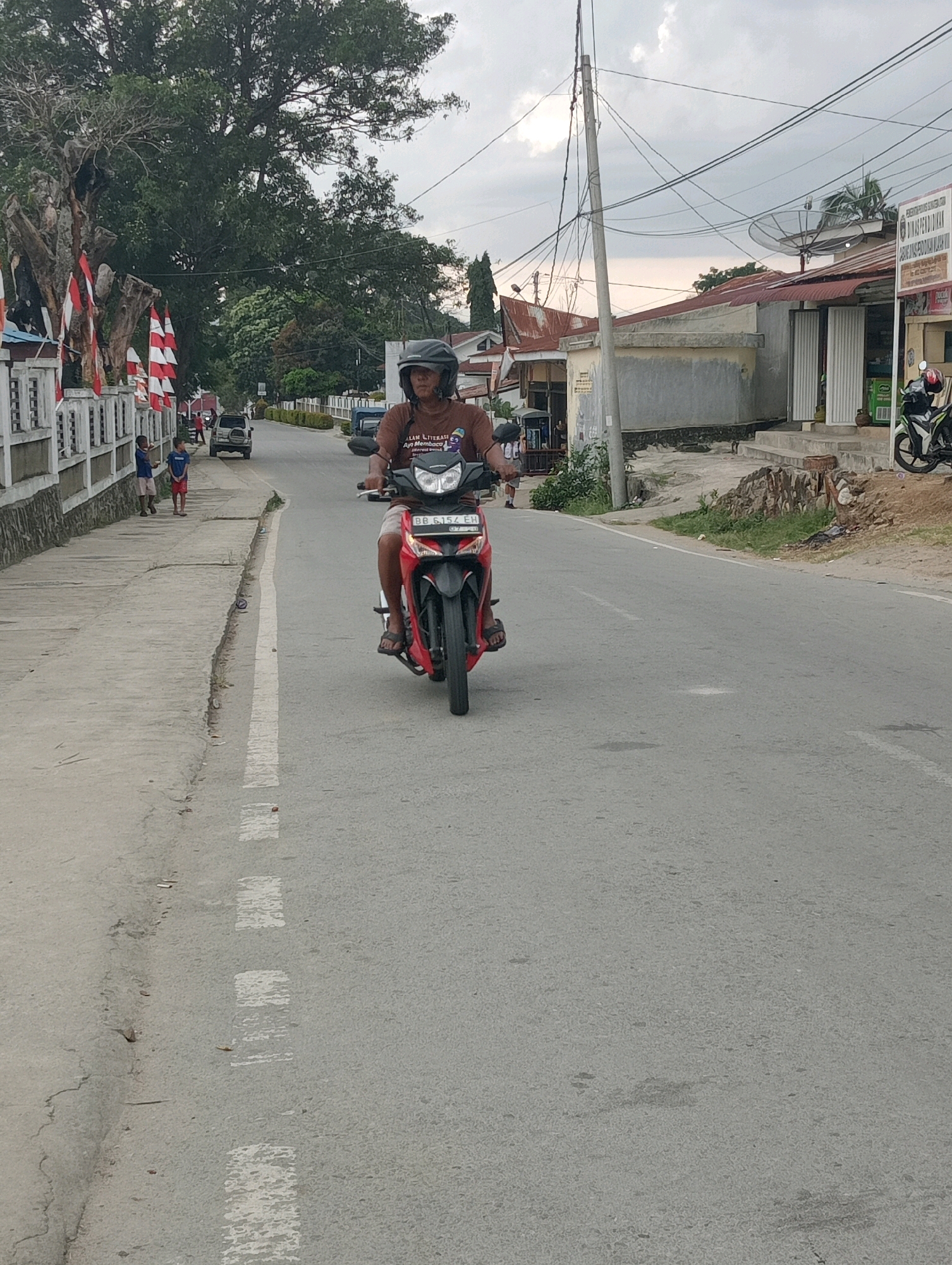 In the city of Hinalangbagasan, Indonesia, a person wearing a helmet rides a red motorcycle down a paved road. The motorcycle has a license plate that reads "BB 6154 EH". The rider is wearing a brown t-shirt with text that reads "ALAM LITERASI AYOO MEMBACA" and a dark helmet. The road has a white dashed line painted on it. On the left side of the road, there is a sidewalk with a white railing adorned with red and white flags. Two young boys are standing on the sidewalk. In the background, there are trees, buildings with red roofs, and a satellite dish on top of one building. The sky is overcast.