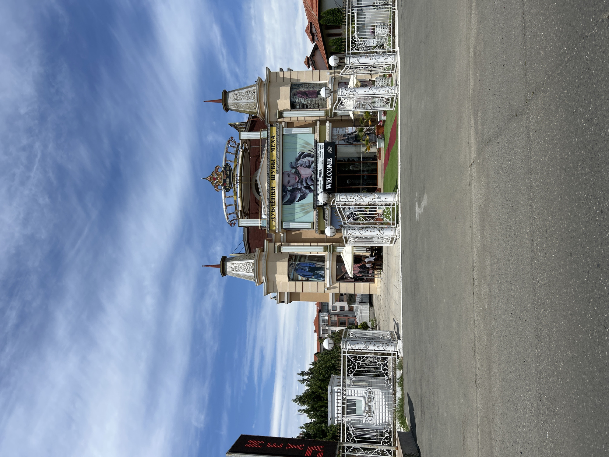 The image captures the exterior of a building in Aheloy, Bulgaria, identified by signage as "KOZHANOTO TSARSTVO" (Leather Kingdom) and "DUBLEANKI, SHUBY, MEHA" (Sheepskin Coats, Fur Coats, Furs). The building has an ornate facade with multiple levels, decorative towers, and large banners displaying images of fashion models and clothing. A prominent sign at the entrance reads "WELCOME" above a red carpet leading into the establishment. White ornate metal gates and fencing surround the property, with a paved area in front. Trees and residential buildings are visible in the background. The sky is blue with scattered clouds.