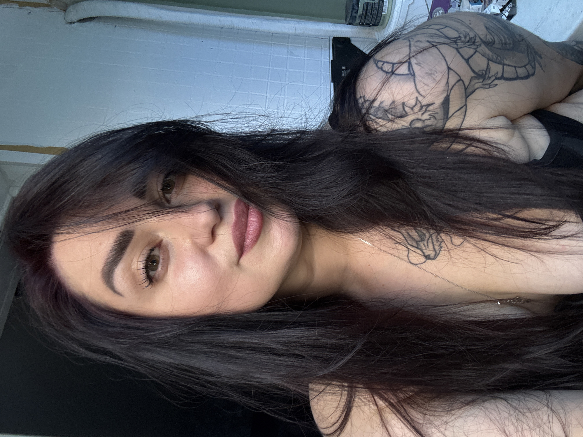 A selfie shows a woman with long, dark hair and brown eyes. She is wearing makeup, including lipstick and mascara. She has a tattoo on her right shoulder and a necklace with a pendant. The background is out of focus and appears to be an indoor setting with a wall and some objects on a shelf. The lighting suggests it is daytime.