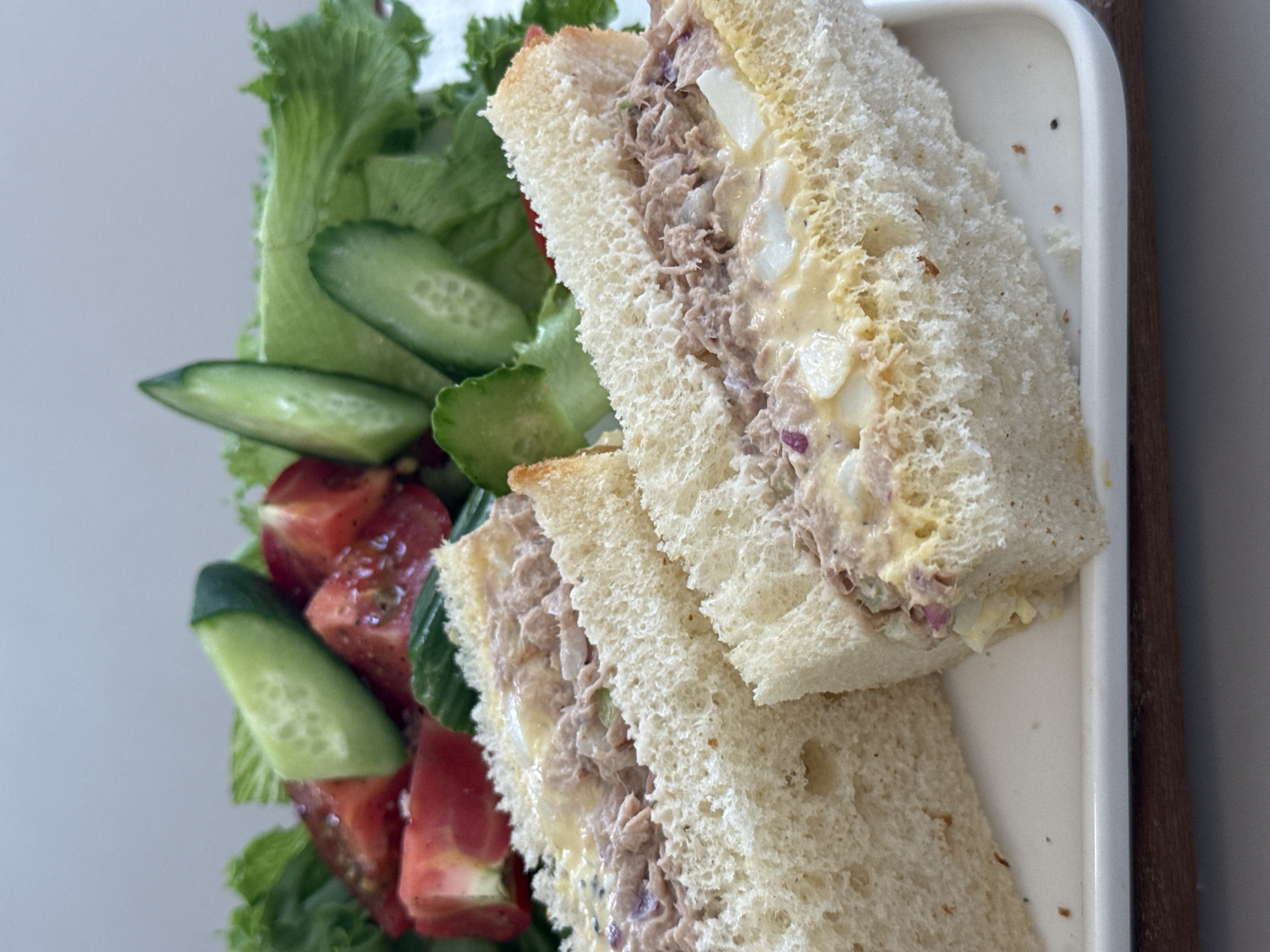 A close-up, high-angle shot captures two halves of a tuna salad sandwich resting on a white rectangular plate. The sandwich is made with white bread, and the filling appears to be a mixture of tuna, mayonnaise, chopped eggs, and red onion. A side salad, composed of lettuce, sliced tomatoes, and cucumber wedges, is visible behind the sandwiches. The background is a plain, light gray wall. The scene is well-lit, highlighting the textures and colors of the food. No people or city elements are visible.