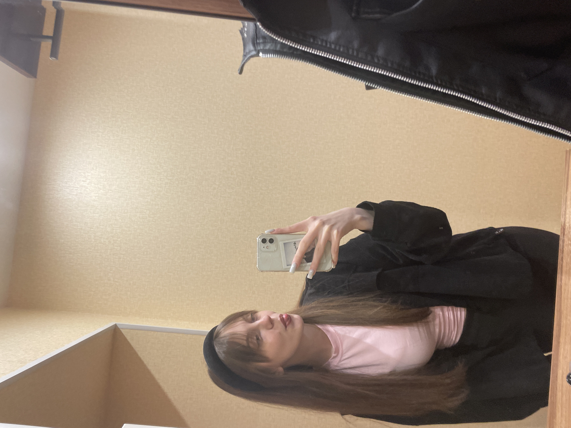 A woman is taking a selfie in a mirror. The background consists of a patterned wall, seemingly inside a closet or dressing room. The woman is wearing a pink shirt with a black jacket. No specific landmarks or locations in the city of Ufa, Russia, are discernible from the image.