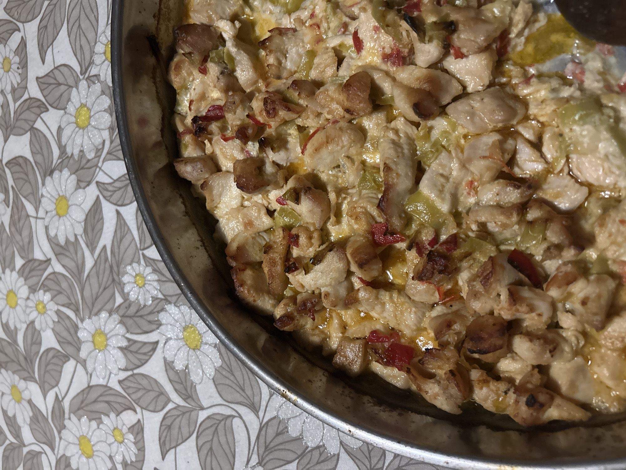 The image shows a close-up, overhead shot of a metal baking dish containing a chicken dish. The chicken appears to be cut into bite-sized pieces and mixed with a creamy sauce, including green and red peppers. The dish is on a tablecloth, featuring a floral pattern of white daisies with yellow centers and gray leaves on a light background. No people are visible in the image. The location of Rakitovo, Bulgaria is extraneous and does not appear to be relevant to the image's contents.