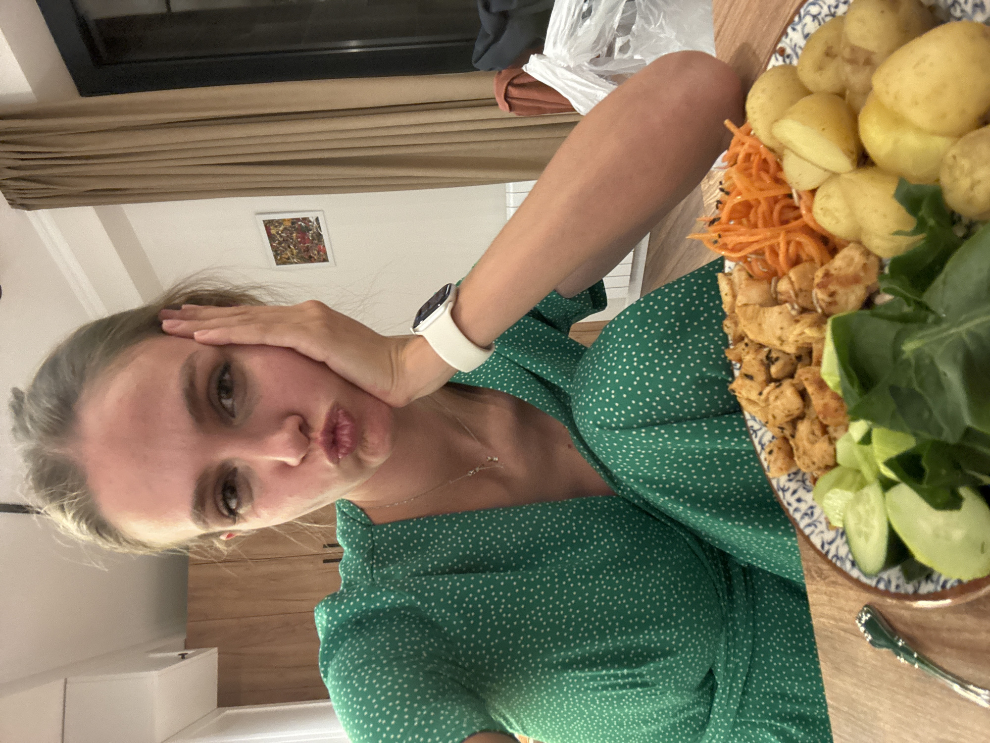 A woman in a green polka-dot dress poses with a plate of food on a wooden table. The plate contains boiled potatoes, shredded carrots, what appears to be diced cooked chicken, and sliced cucumbers with green leafy vegetables. The woman has her hand on her cheek and is making a kissing face towards the camera. She is wearing a watch with a white band. The background includes a window with closed brown curtains, a framed painting on the wall, and kitchen cabinets. The location is inside a home. The city and region are unknown.