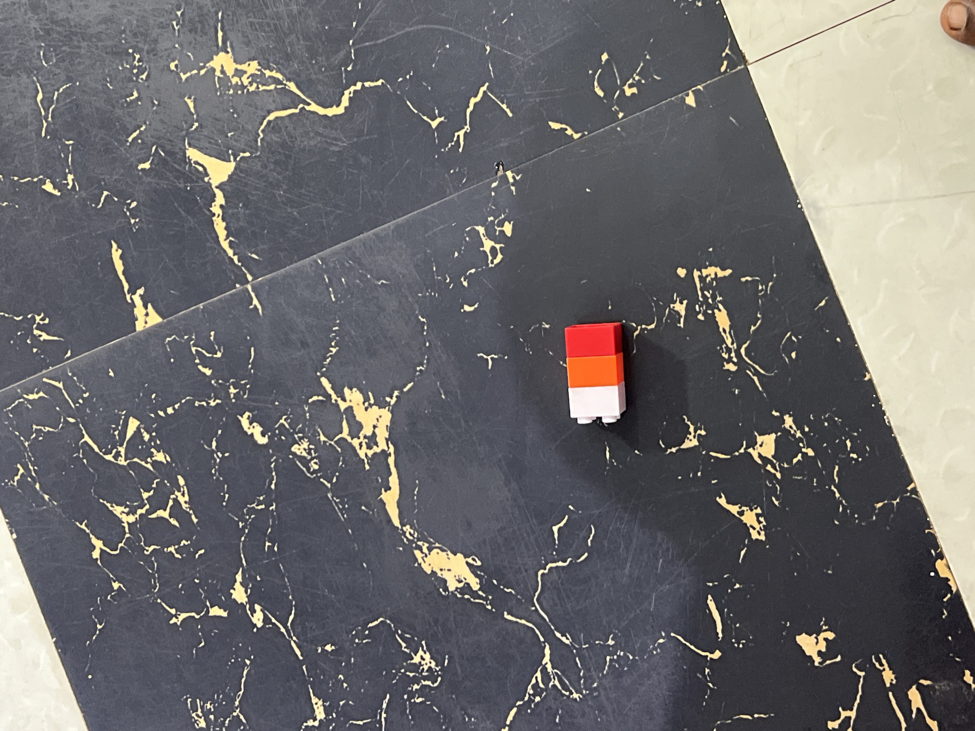 A close-up, top-down shot captures a small, stacked object of three Lego bricks: red on top, then orange, then white. The Lego bricks are situated on a dark, marbled surface with gold veining. A sliver of a lighter, cream-colored tiled surface is visible to the right of the dark surface. A person's thumb and forefinger are visible in the upper right corner, hinting at a hand reaching into the frame, possibly to interact with the Lego bricks. The lighting appears to be artificial or from an overcast sky, creating soft shadows. The scene is likely indoors, given the tiled surfaces. There is no visible text in the image. The overall impression is of a simple, everyday moment, possibly involving a child or someone playing with Lego. The specific location context is Gura Top, Nigeria.