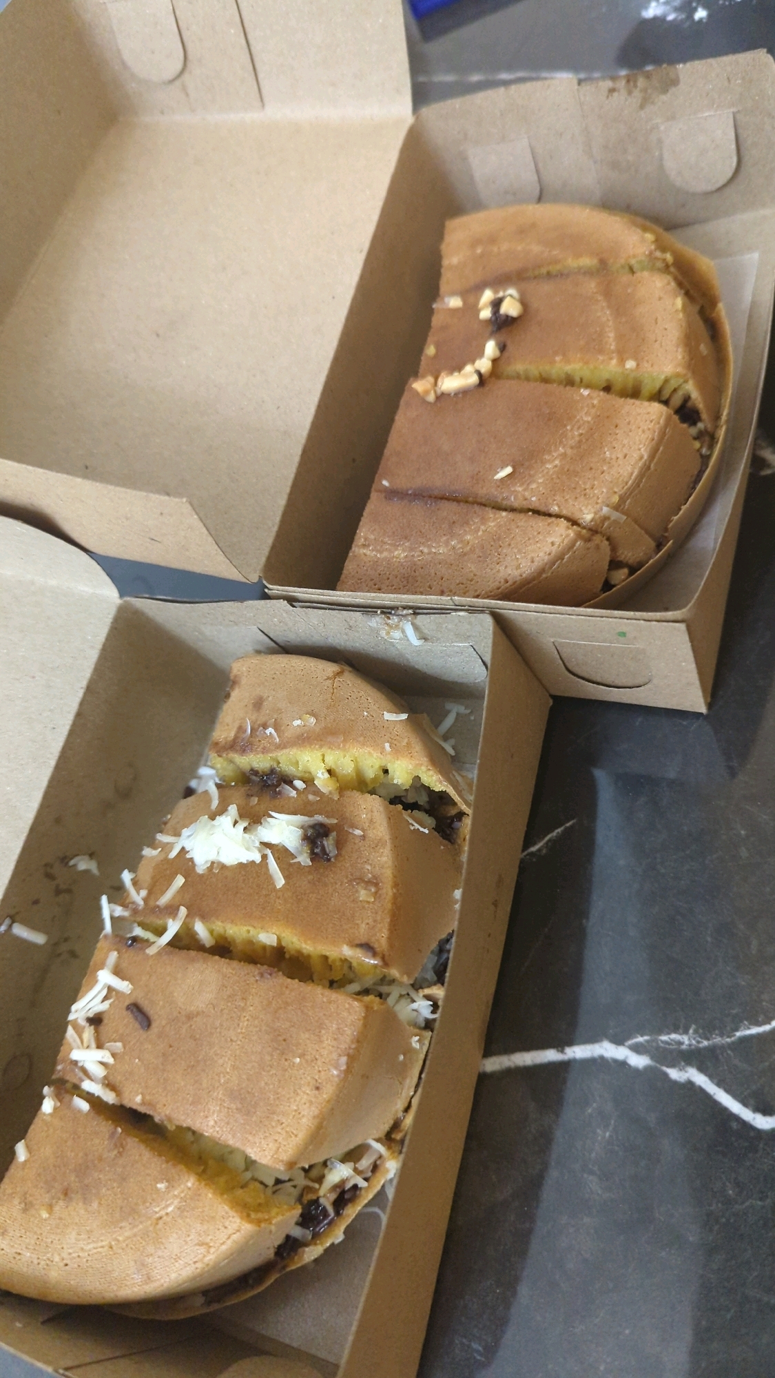Two cardboard take-out boxes, each containing a portion of sweet martabak, are arranged on a dark marble-patterned surface. The martabak are cut into rectangular slices and appear to be filled with chocolate sprinkles and grated cheese. The box in the foreground is closer to the viewer and contains a larger portion of martabak, while the box in the background contains a smaller portion. The lighting suggests an indoor setting, possibly a street food stall in Semut, Indonesia, given the presence of martabak.