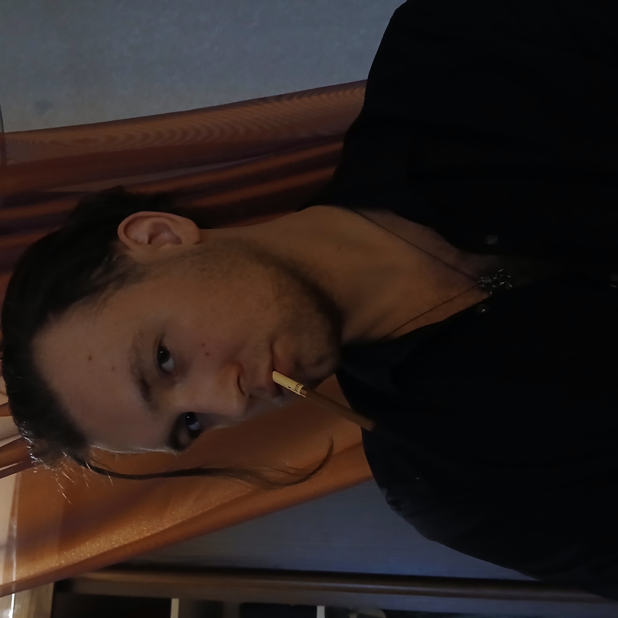The image is a rotated close-up portrait of a man, observed as if lying down or reclining. He has dark hair, styled in a ponytail or bun, with visible stubble on his jawline and chin. His dark, focused eyes are directed straight at the viewer, conveying a neutral or slightly contemplative expression. He is dressed in a dark-colored top, and a metallic, possibly silver, pendant on a chain is visible against his chest. An object, resembling a pen or pencil with a light-colored tip, is held between his lips; part of the word "ping" is discernible on its side.
The setting appears to be indoors, dimly lit. A translucent, reddish-brown fabric, possibly a curtain or drapery, stretches across the foreground and background, creating a soft, warm hue throughout the scene. Behind this fabric, a muted blue-gray wall or surface is faintly visible, particularly in the upper portion of the frame. The combined effect of the lighting and warm tones evokes an intimate and somewhat subdued atmosphere. This scene is contextualized as being in Yekaterinburg, Russia.