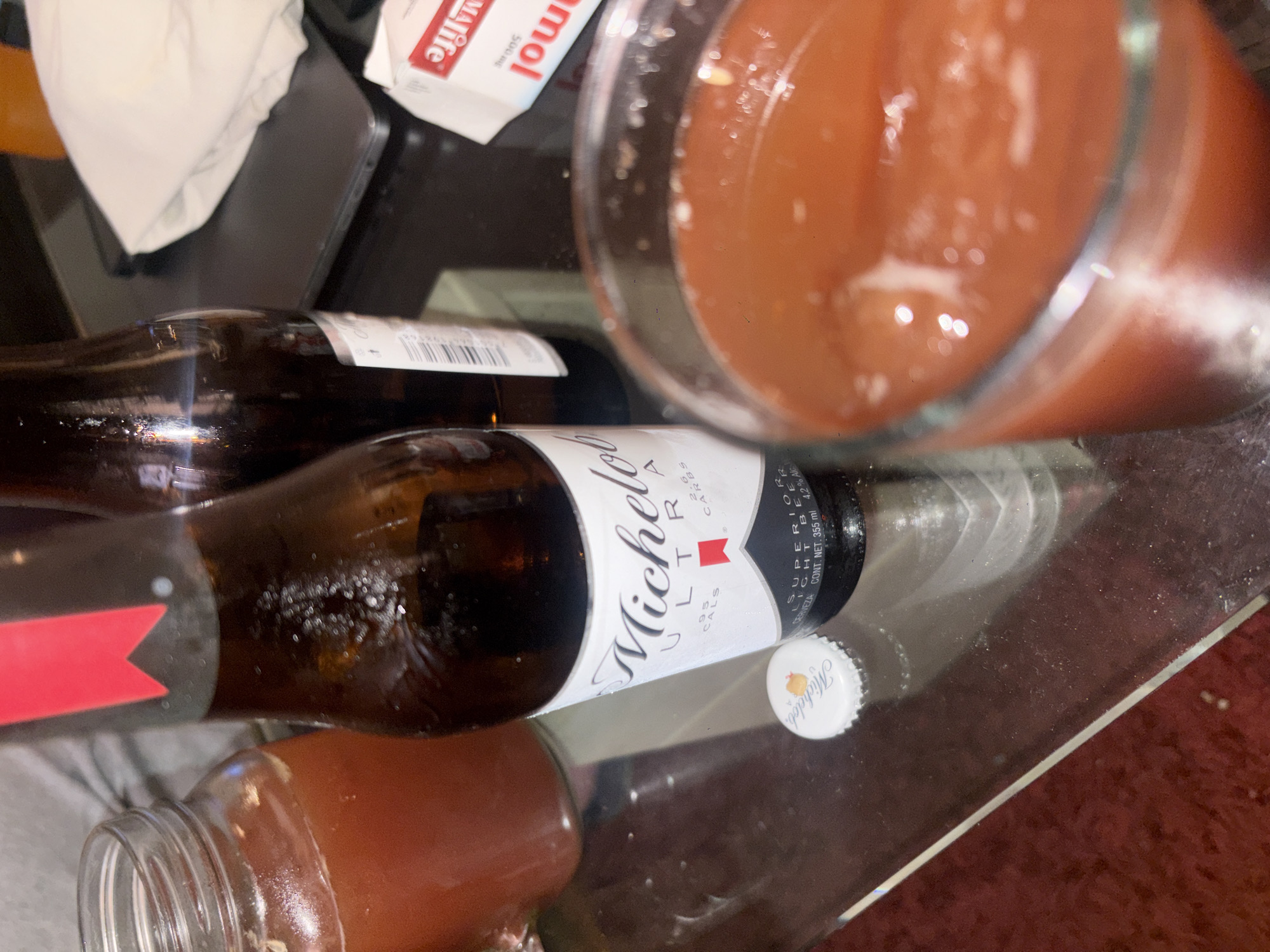 A cluttered tabletop scene includes a Michelob Ultra beer bottle lying horizontally, its white label prominently displaying the brand name. Nearby, a glass is filled with a reddish-brown liquid, possibly a mixed drink or tomato juice. A white pill box is also visible, partially obscured by other objects. The background is blurred, indicating a shallow depth of field, and the tabletop surface appears to be reflective, possibly glass. There are no people visible in the frame. It is not possible to know if this is in Mexico City, Mexico.