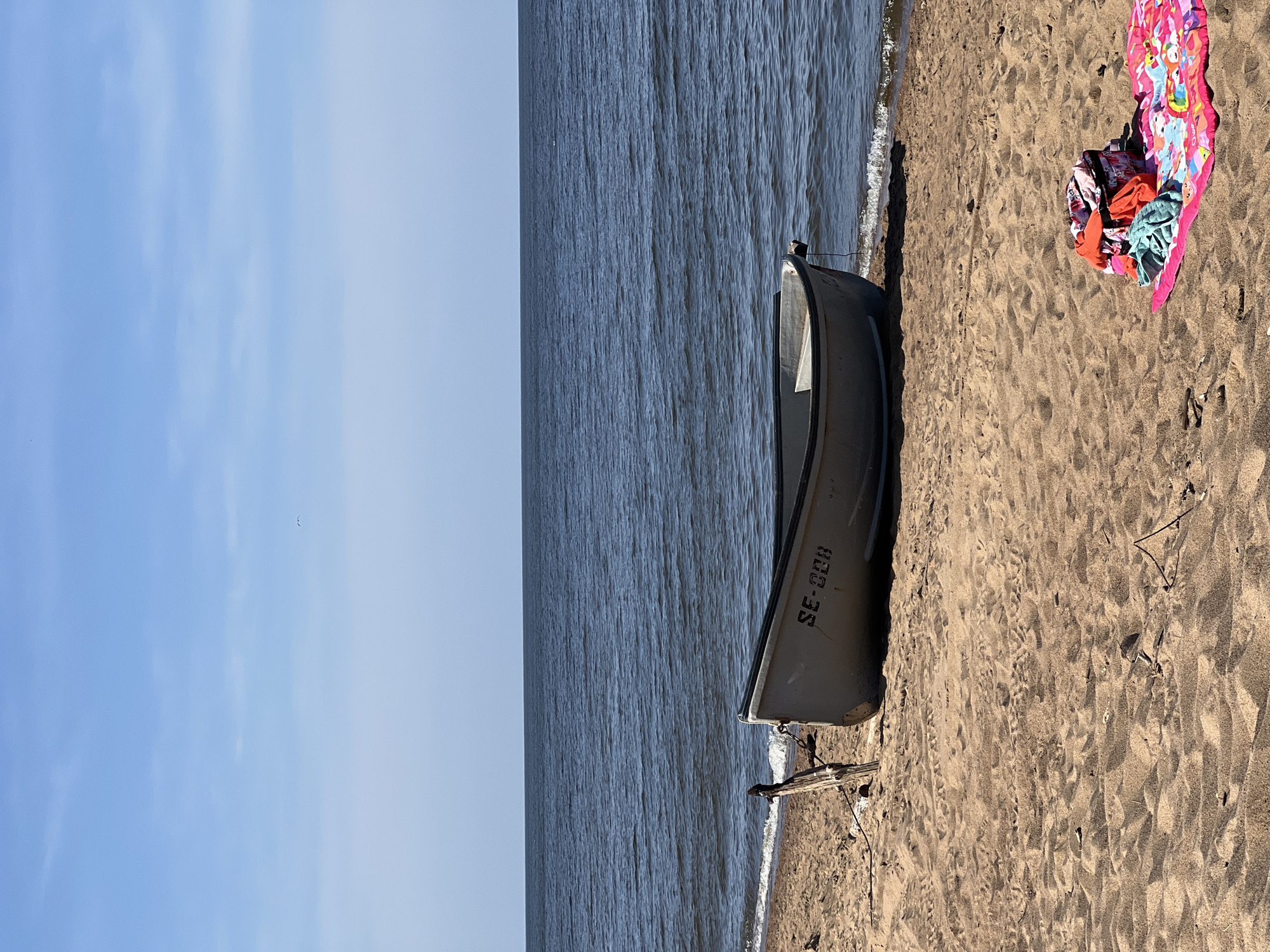 A light gray rowboat with the registration SE-CCH is pulled up on a sandy beach. The boat is positioned in the foreground to the left of a pile of colorful beach towels and bags. The ocean is visible in the background, with gentle waves lapping at the shore under a clear blue sky. A single dark bird is visible in flight against the sky.