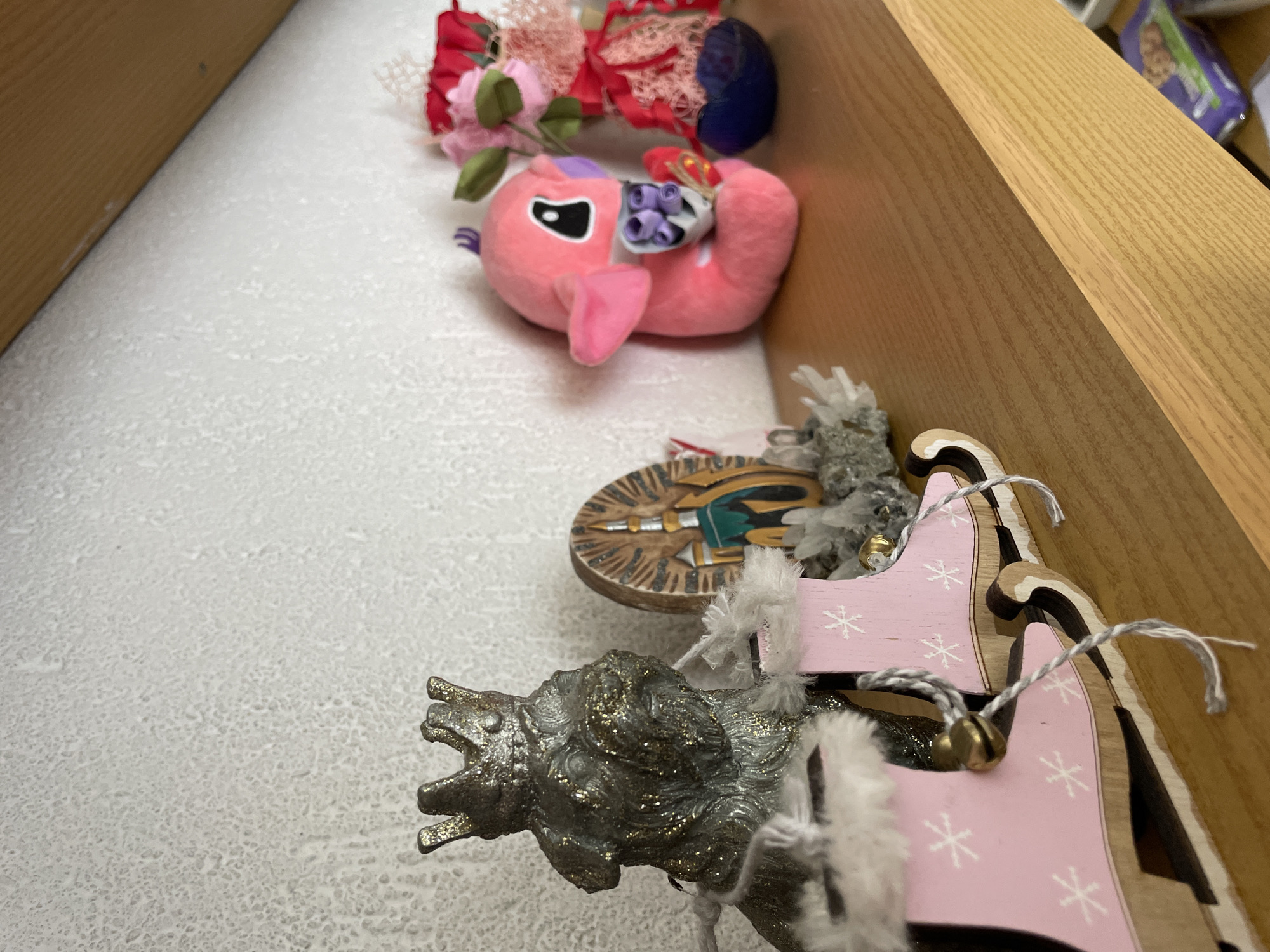 The image displays a shelf with various decorative items, positioned against a textured white wall. On the left side of the shelf, a small, metallic figurine of a dog wearing a crown is visible, adorned with gold glitter. In front of the dog figurine, a pair of miniature pink wooden sleighs with white snowflake designs and faux fur trim are placed. Each sleigh has a small bell attached. To the right of the dog figurine, a circular wooden ornament featuring a depiction of a building with radiating lines is present. Further to the right, a pink plush toy, resembling a character from a cartoon with large eyes and floppy ears, is holding a small bunch of purple flowers. Behind the pink toy, a decorative arrangement of pink and red artificial flowers in a blue vase is visible. The shelf itself is made of light brown wood, and a portion of the ceiling, which is angled and also appears to be wood, is visible in the upper right corner. The overall setting suggests a home interior or a shop display, likely in the city of Zlatograd, Bulgaria, given the context provided.