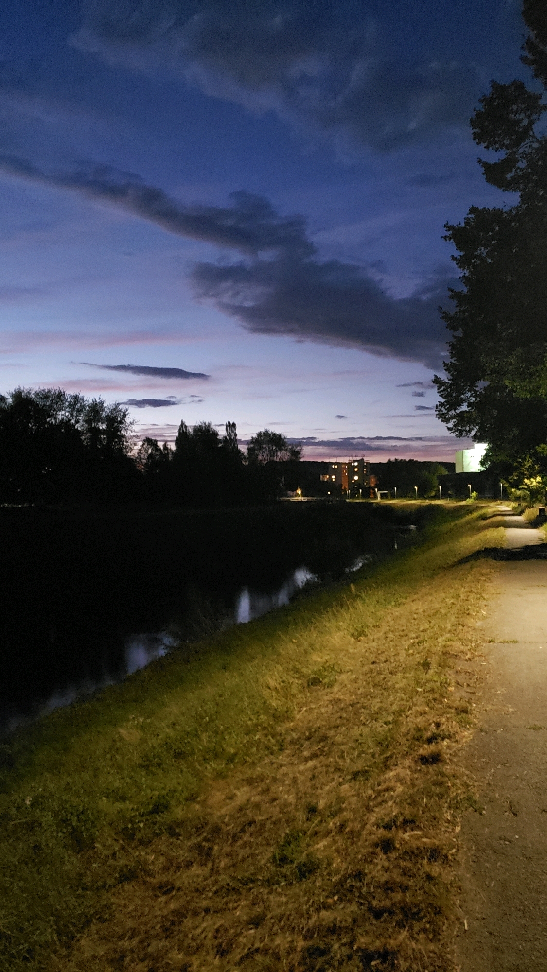The image depicts a dusky scene along a paved path bordering a body of water. The sky above is a deep blue, transitioning to lighter hues near the horizon, with streaks of pink and purple, and scattered dark clouds. On the left side of the image, silhouetted trees and foliage line the bank of the dark water, which reflects the muted colors of the sky. Across the water, a building with illuminated windows is visible, set against a line of trees. The path on the right is well-lit by a streetlamp, casting a warm glow on the dry, yellowish grass and the pavement. A large, dark tree looms in the upper right corner of the frame, its branches reaching towards the sky. The scene suggests an urban park or waterfront area in Prešov, Slovakia, during twilight. There are no people visible in the image.